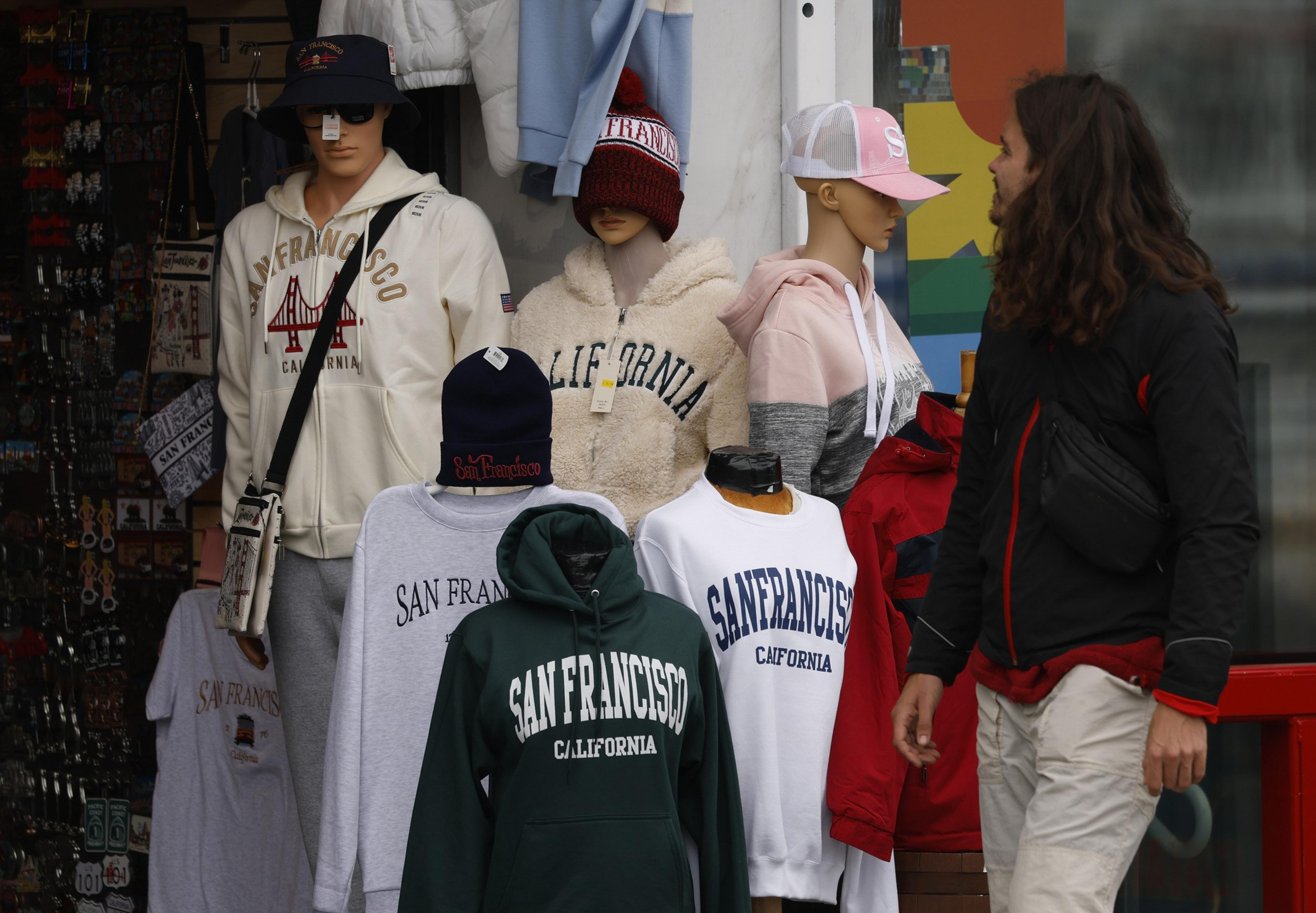The image shows mannequins displaying San Francisco-themed hoodies and shirts. A person with long hair walks by, wearing a black and red jacket.