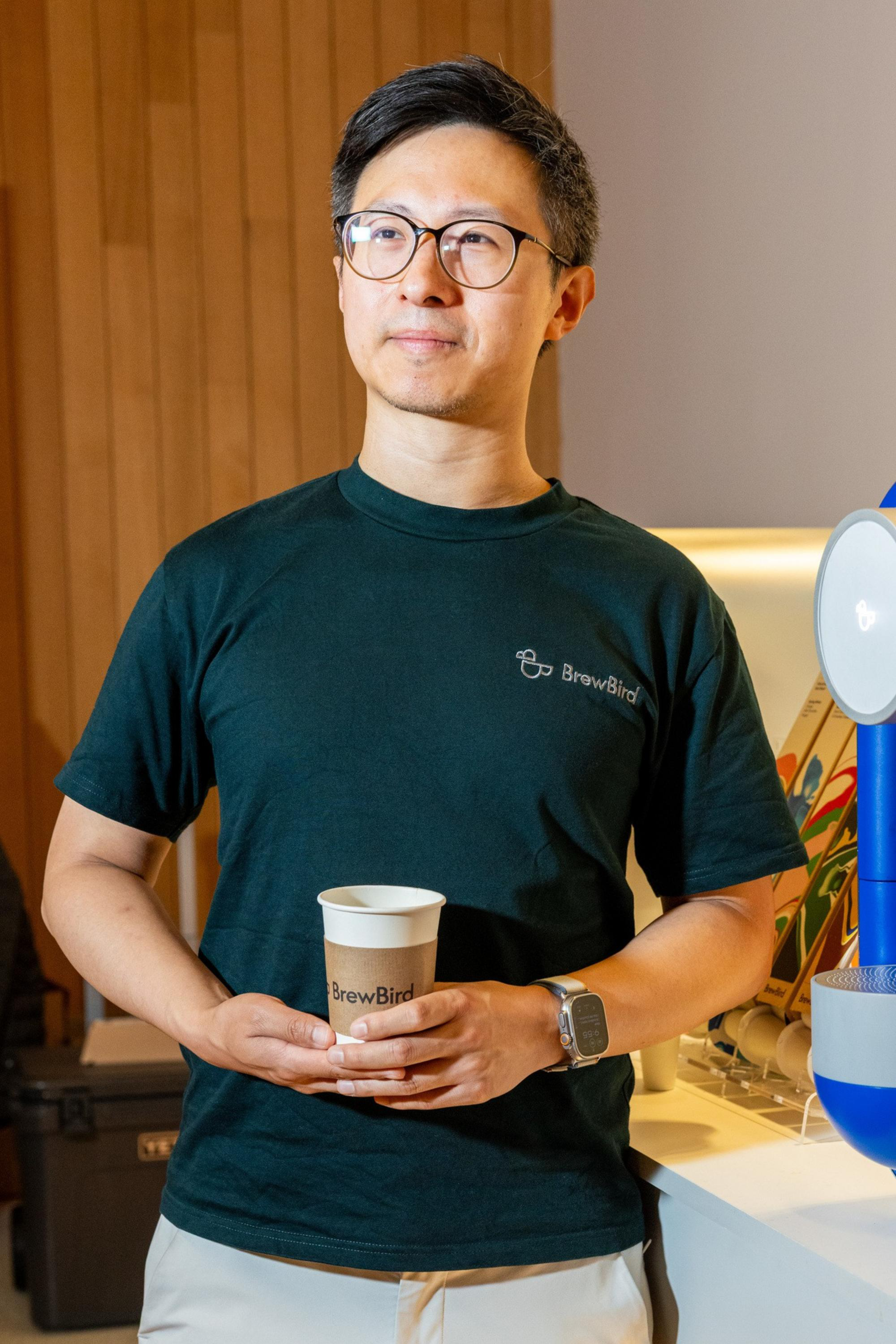 A man wearing glasses and a dark green “BrewBird” t-shirt holds a paper cup, standing next to a coffee machine on a counter.