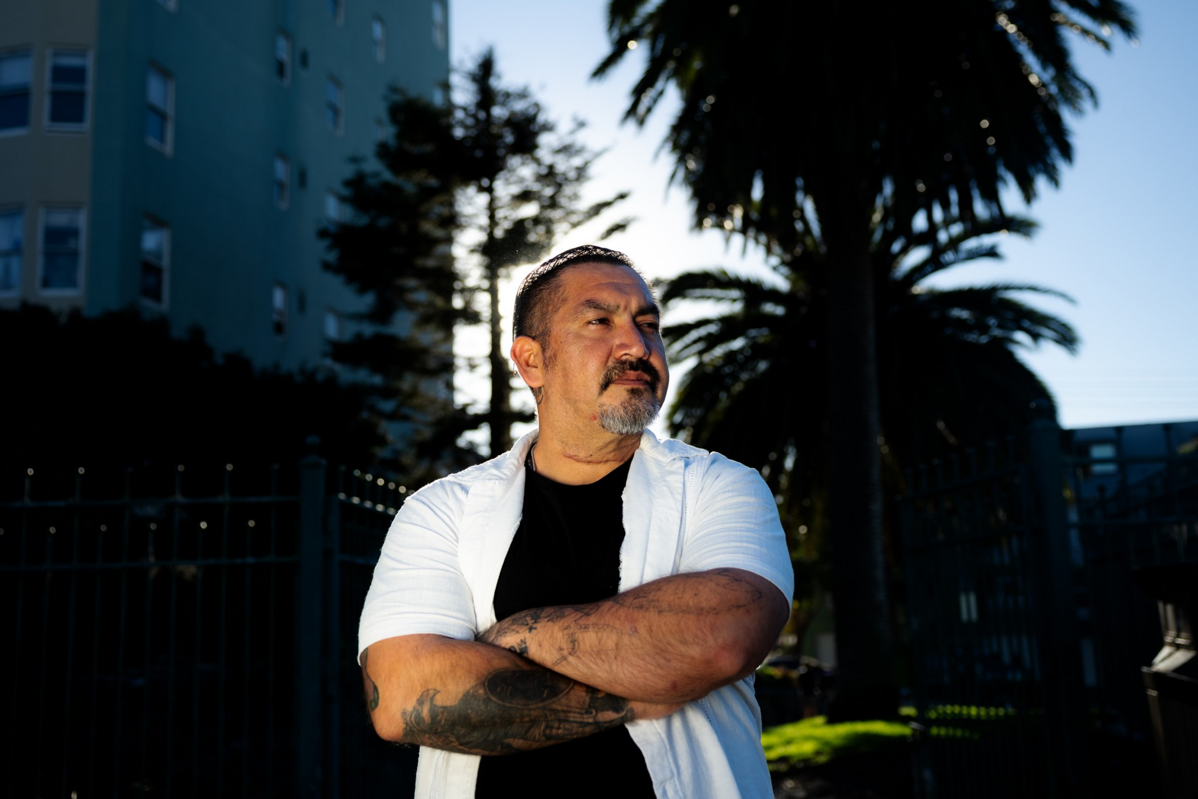 A man with tattoos and a short beard stands with arms crossed, wearing a white short-sleeve shirt over a black t-shirt, with palm trees and buildings behind him