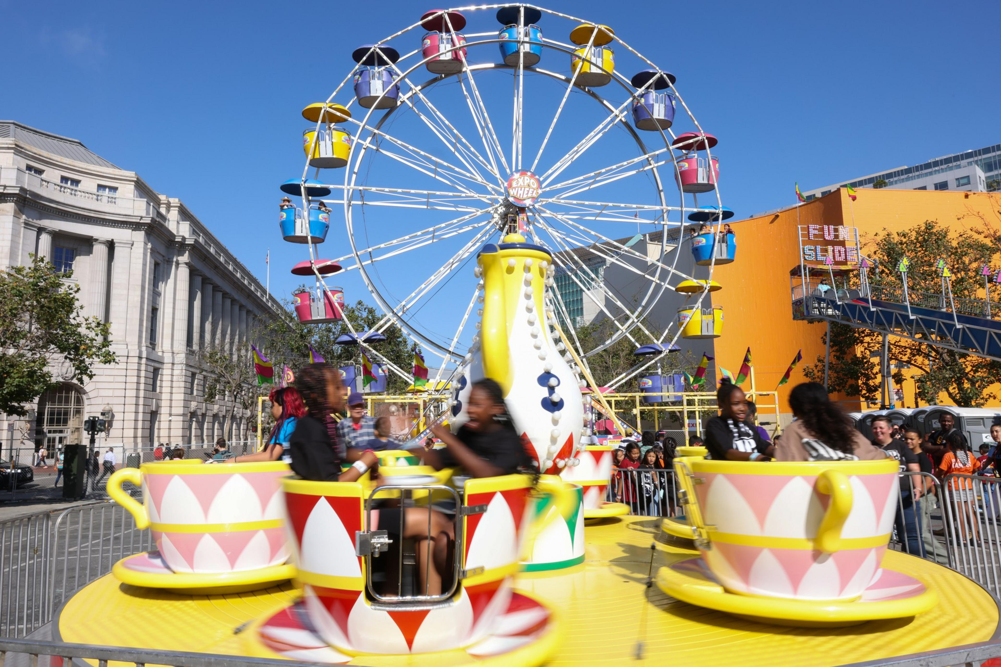 Children ride colorful spinning teacups in front of a large Ferris wheel at an outdoor fair on a sunny day.