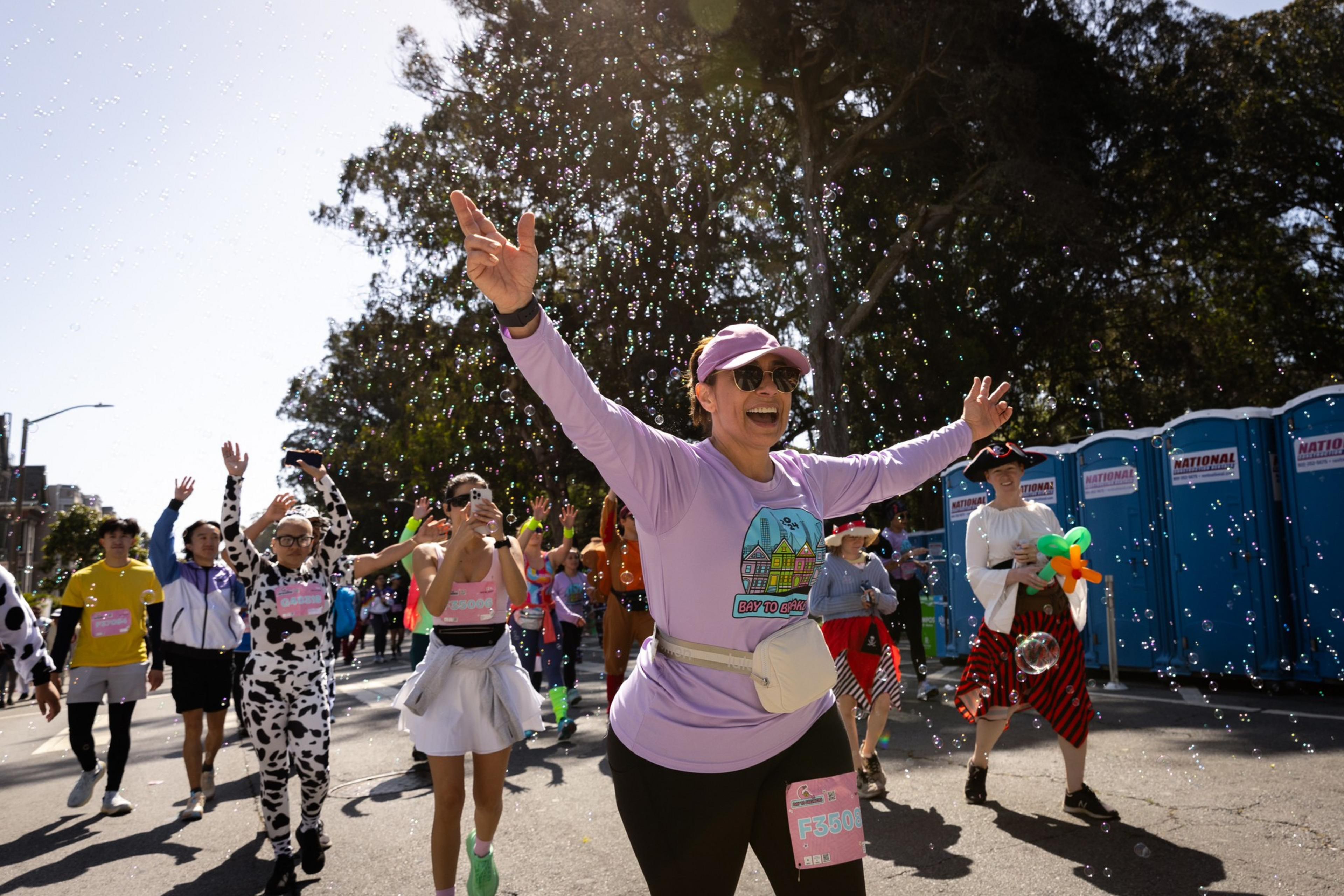 A joyful crowd participates in a festive event under a sunny sky, surrounded by bubbles with participants in colorful costumes, smiling broadly.
