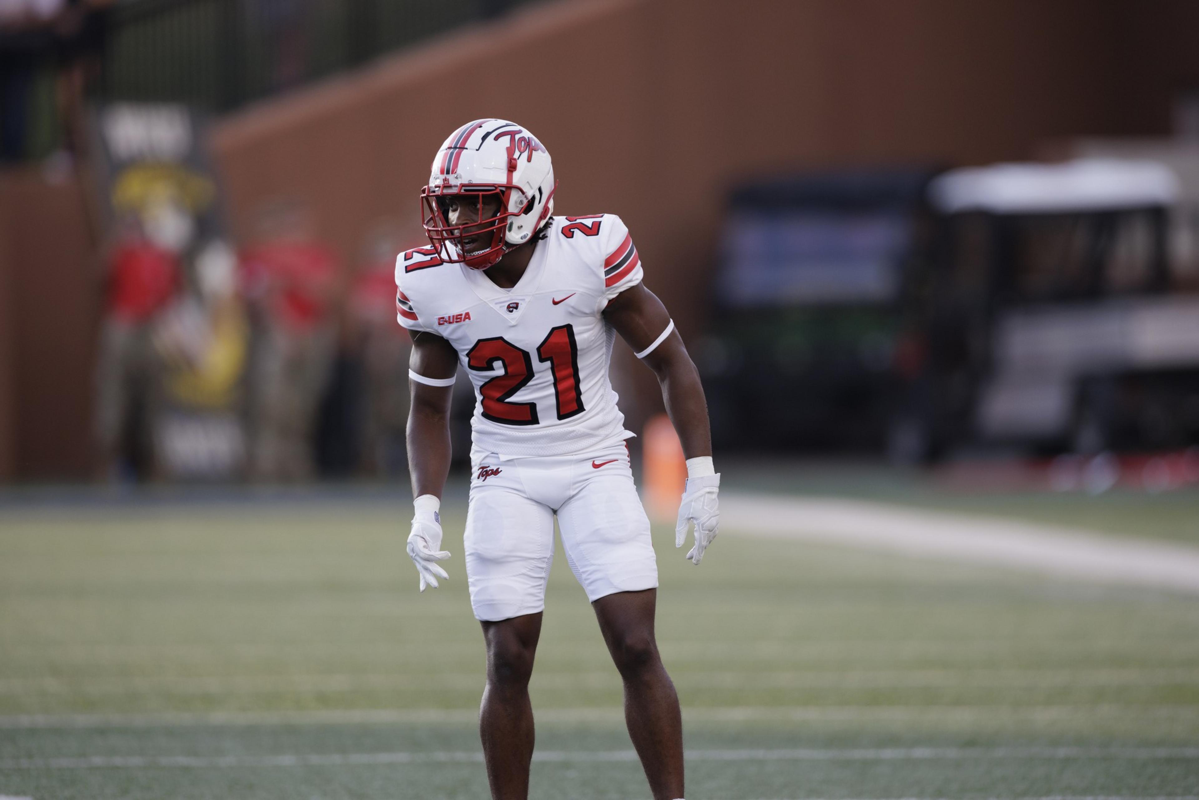 A football player in a white and red uniform with the number 21 prepares on the field. He looks focused, wearing a helmet and gloves, with a blurred background.