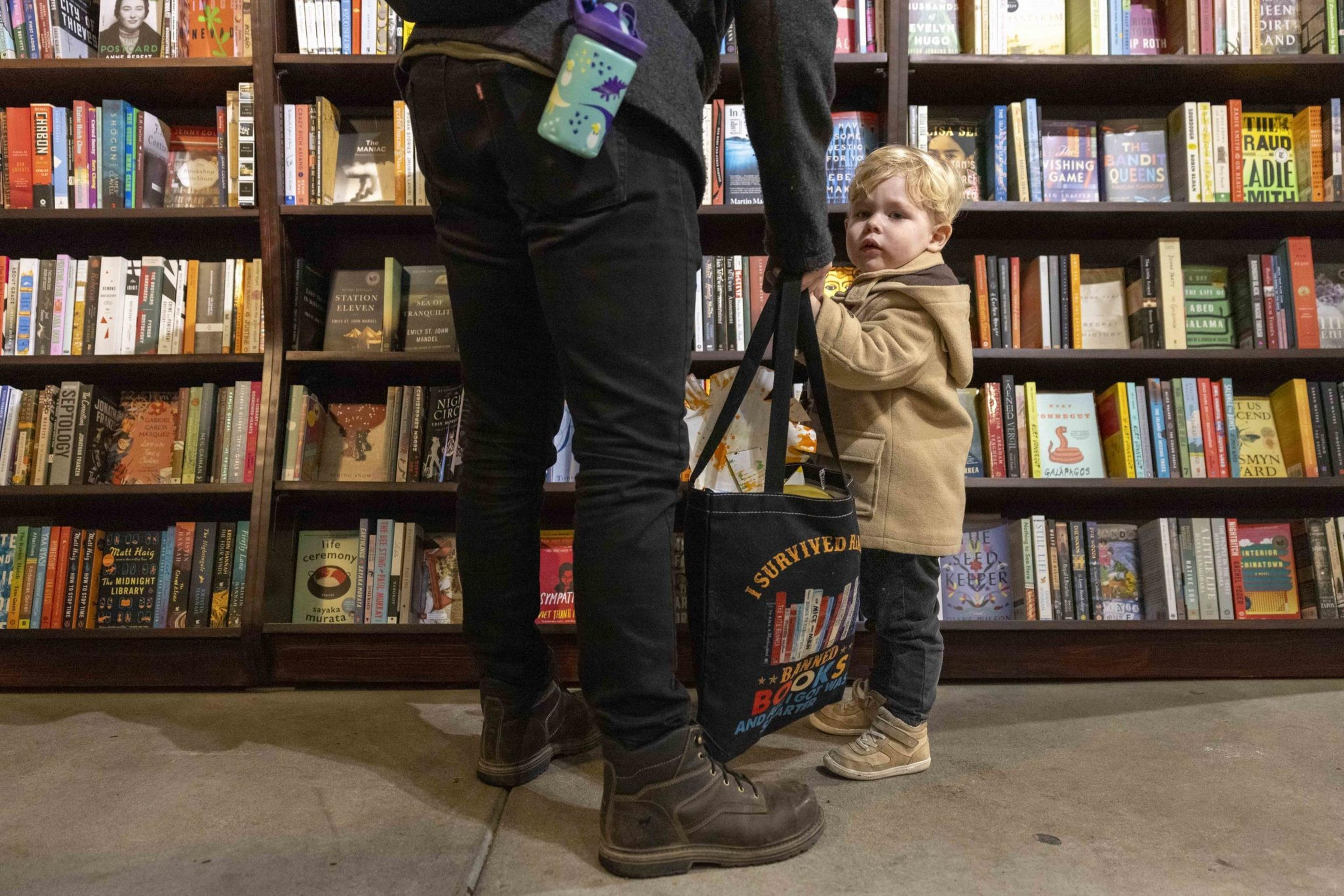 A child in a tan coat stands next to an adult holding a shopping bag in a bookstore. The shelves are full of various colorful books. The child looks curious.