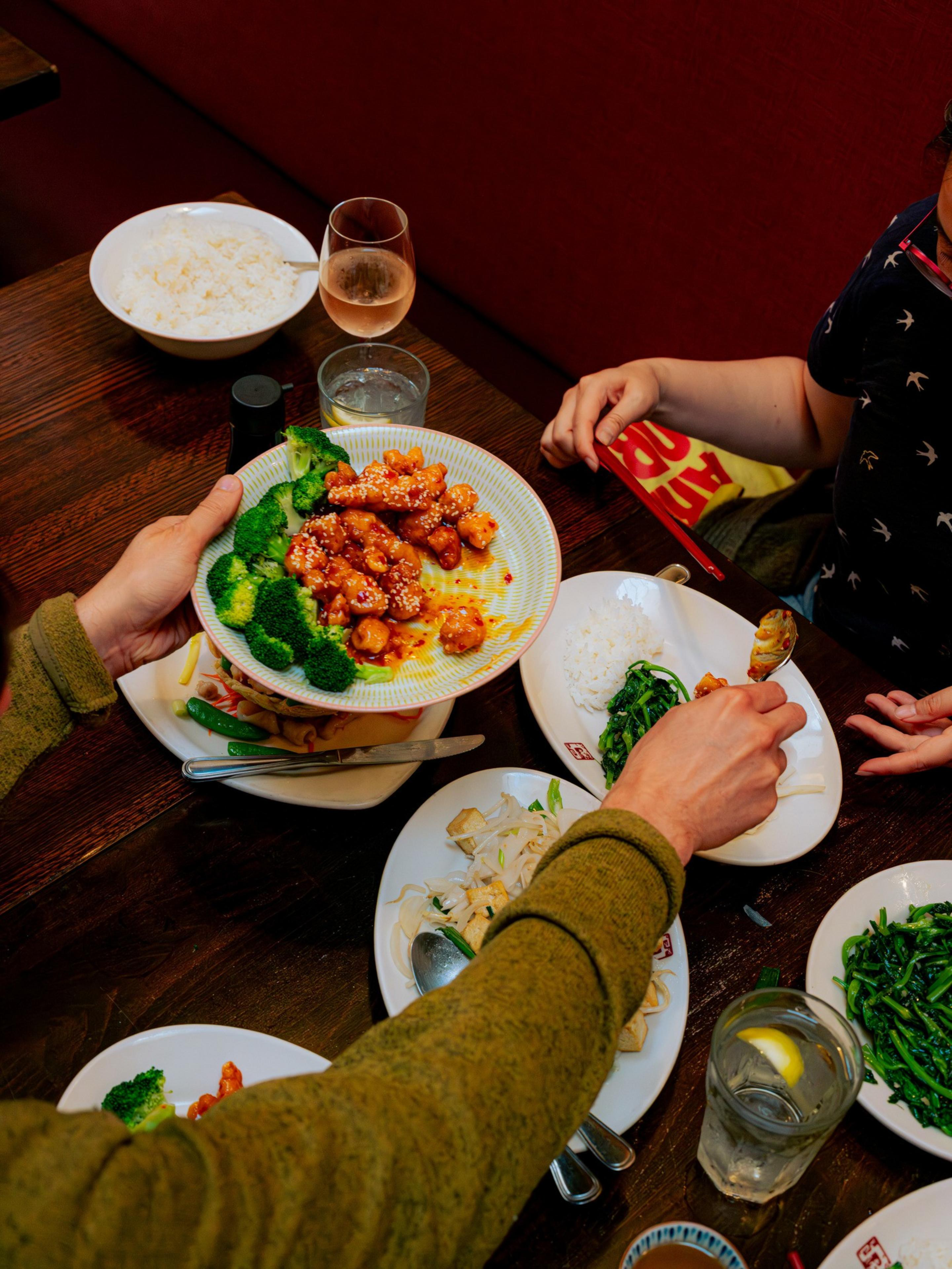 A table with people enjoying a meal featuring broccoli, sesame-covered chicken, rice, stir-fried greens, with drinks including a glass of water with lemon and a glass of wine.