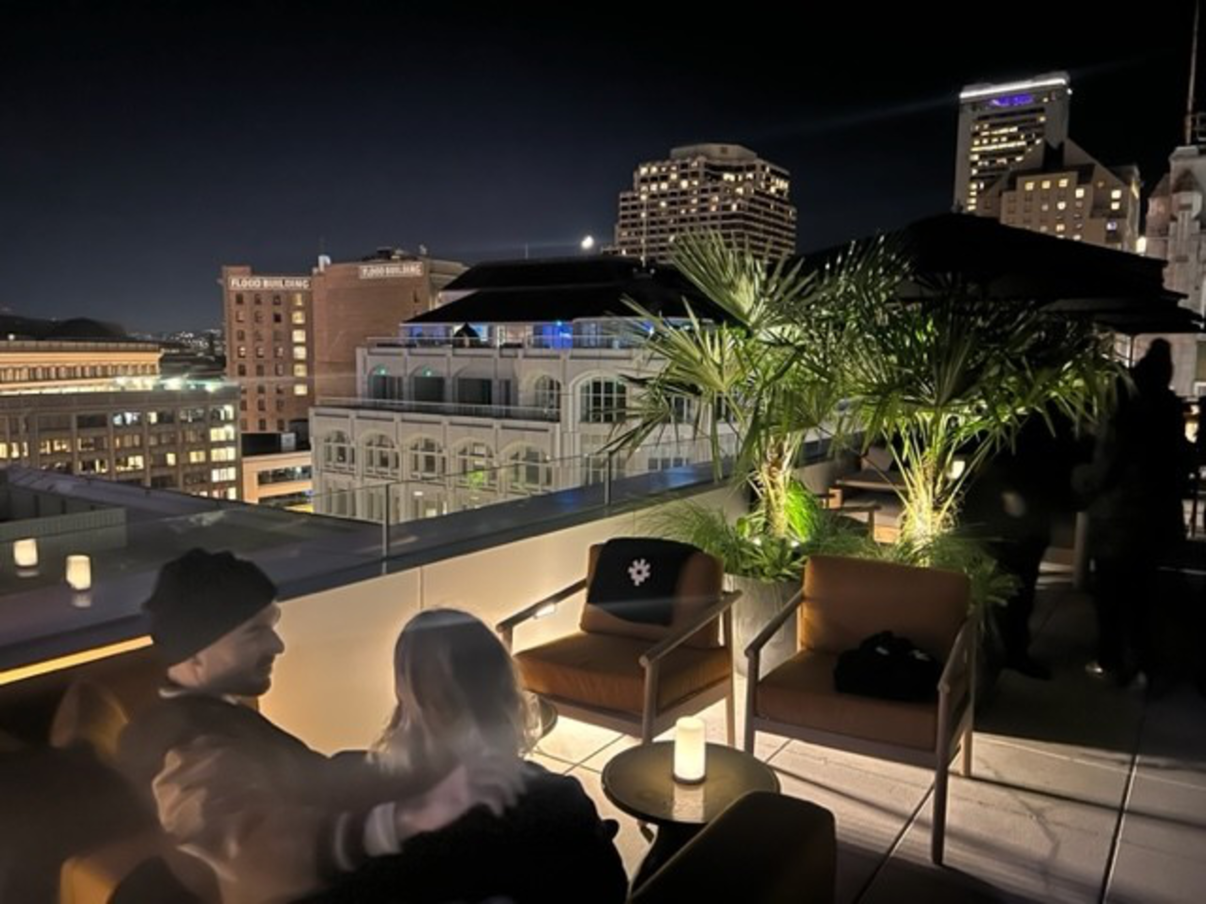 A rooftop lounge at night with cozy seating, soft candlelight, potted plants, and city buildings illuminated in the background.