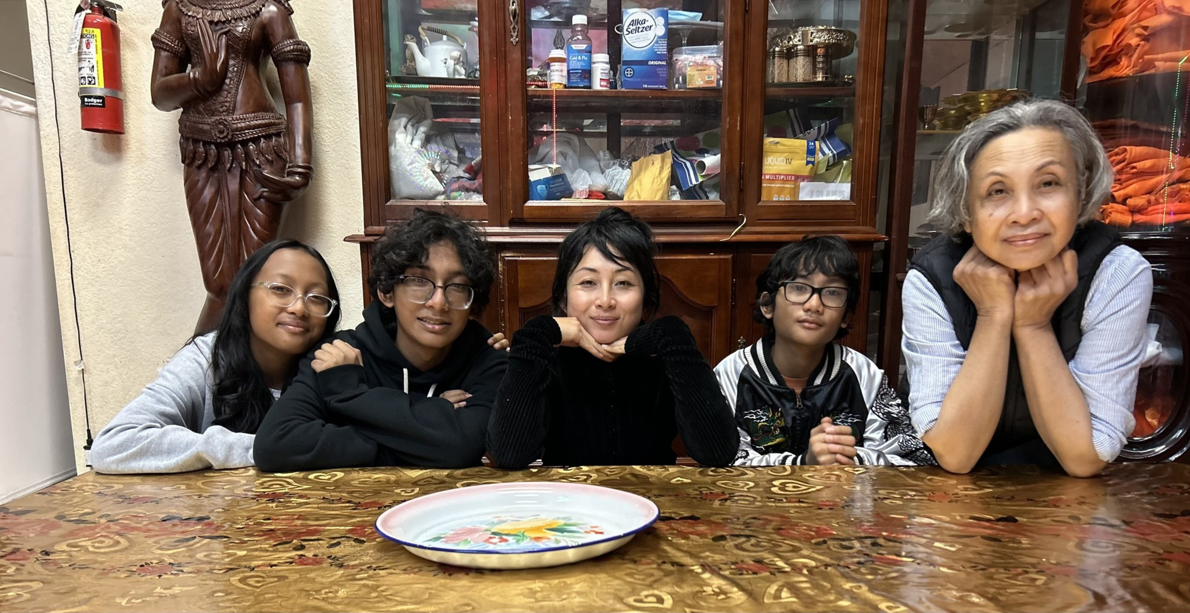 Five people sit closely together behind a decorated wooden table, smiling warmly, with a cabinet and statue in the background.