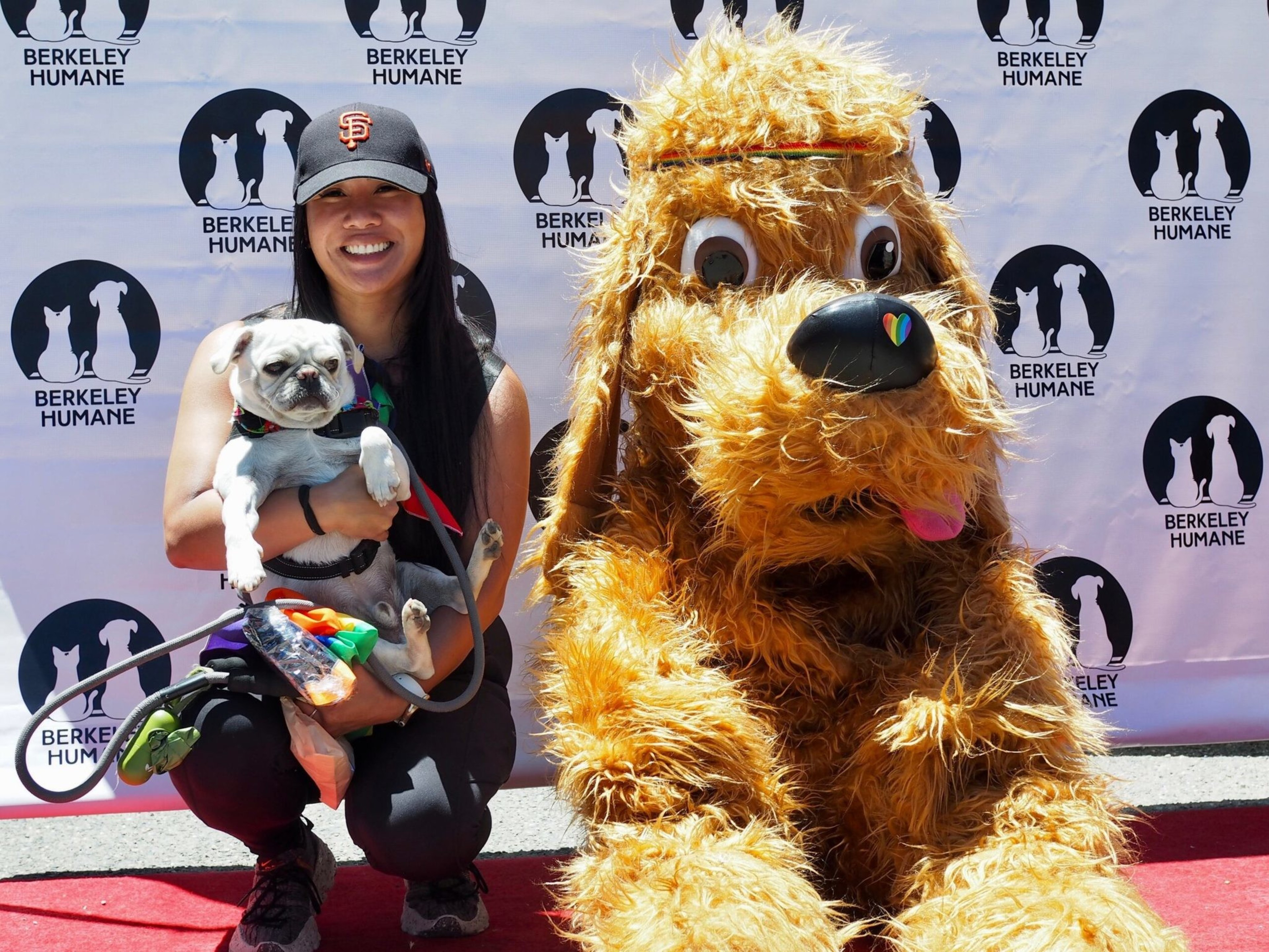 A smiling woman in a San Francisco Giants cap holds a pug, while a large furry dog mascot with a rainbow heart nose sits beside her on a red carpet.