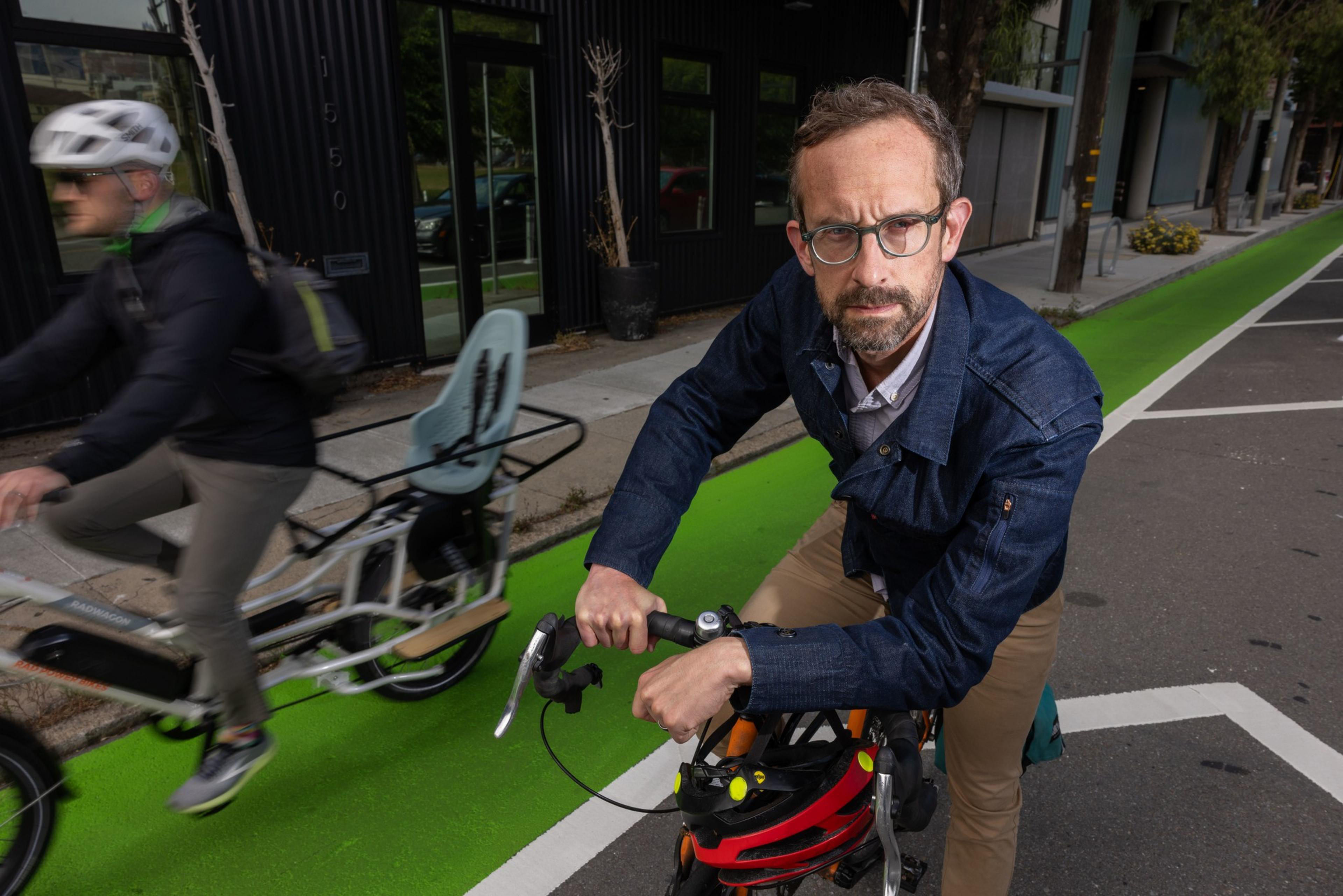A man with glasses and a serious expression stands with his bike on a green bike lane, while another cyclist, in motion, passes by on the left.