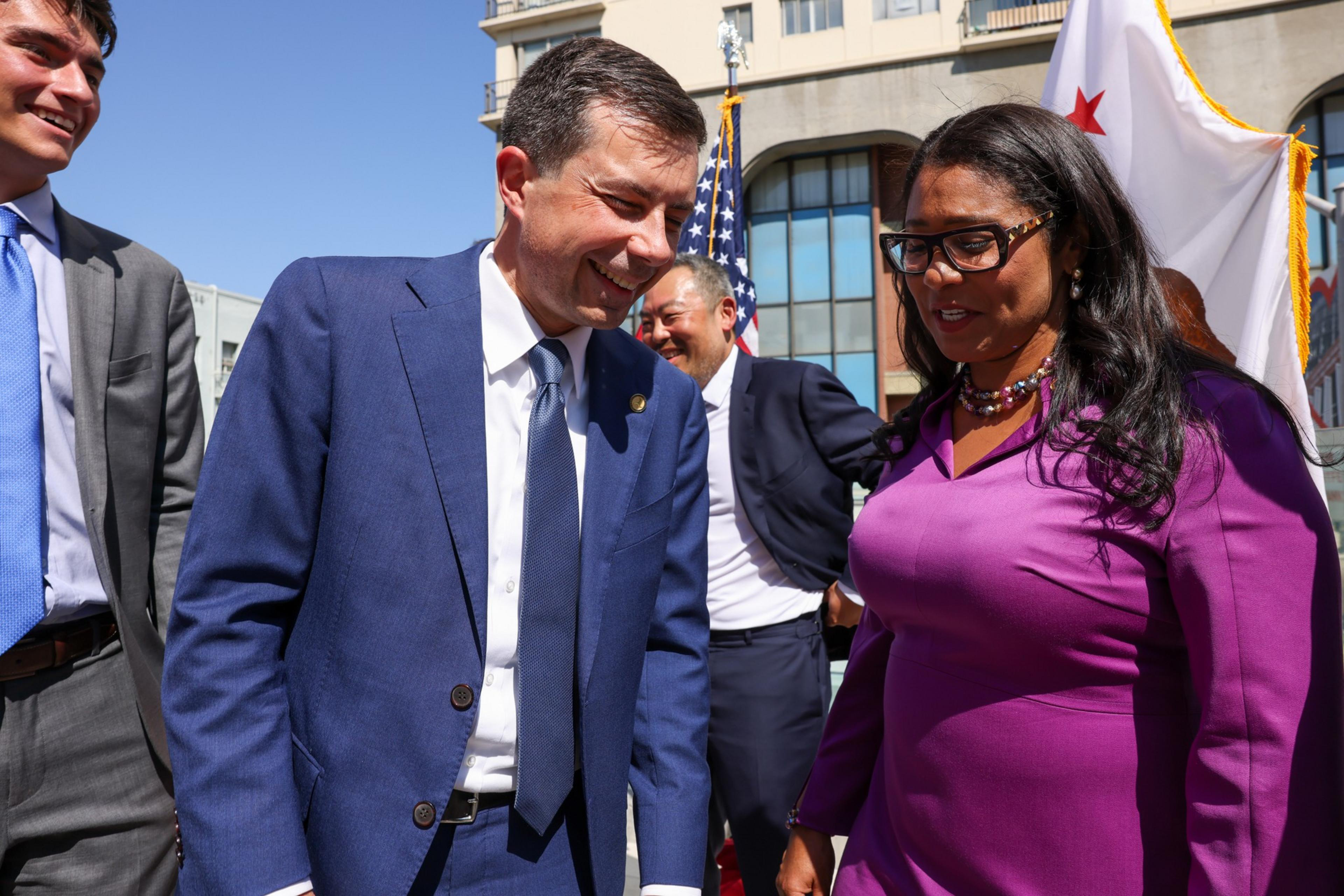 A man in a blue suit smiles while talking to a woman in a purple dress wearing glasses; two other men in suits stand nearby outdoors with flags behind them.