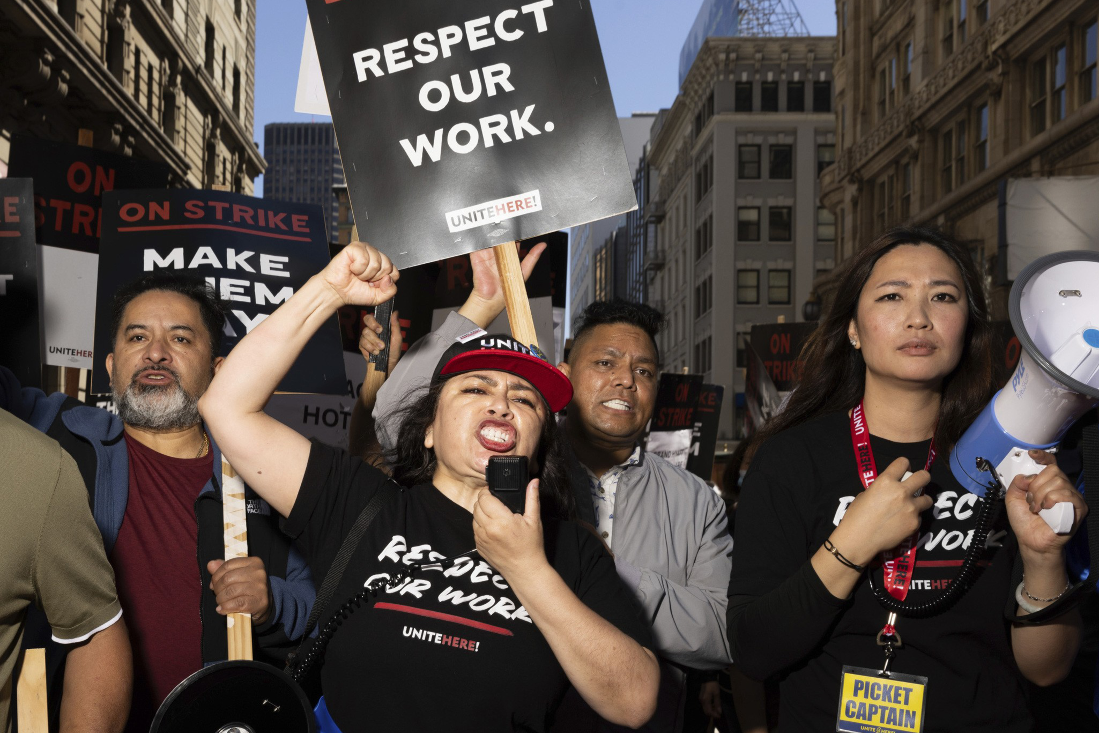 A group of people are passionately protesting in a city street, holding signs that read "RESPECT OUR WORK" and "ON STRIKE." One person speaks into a megaphone.