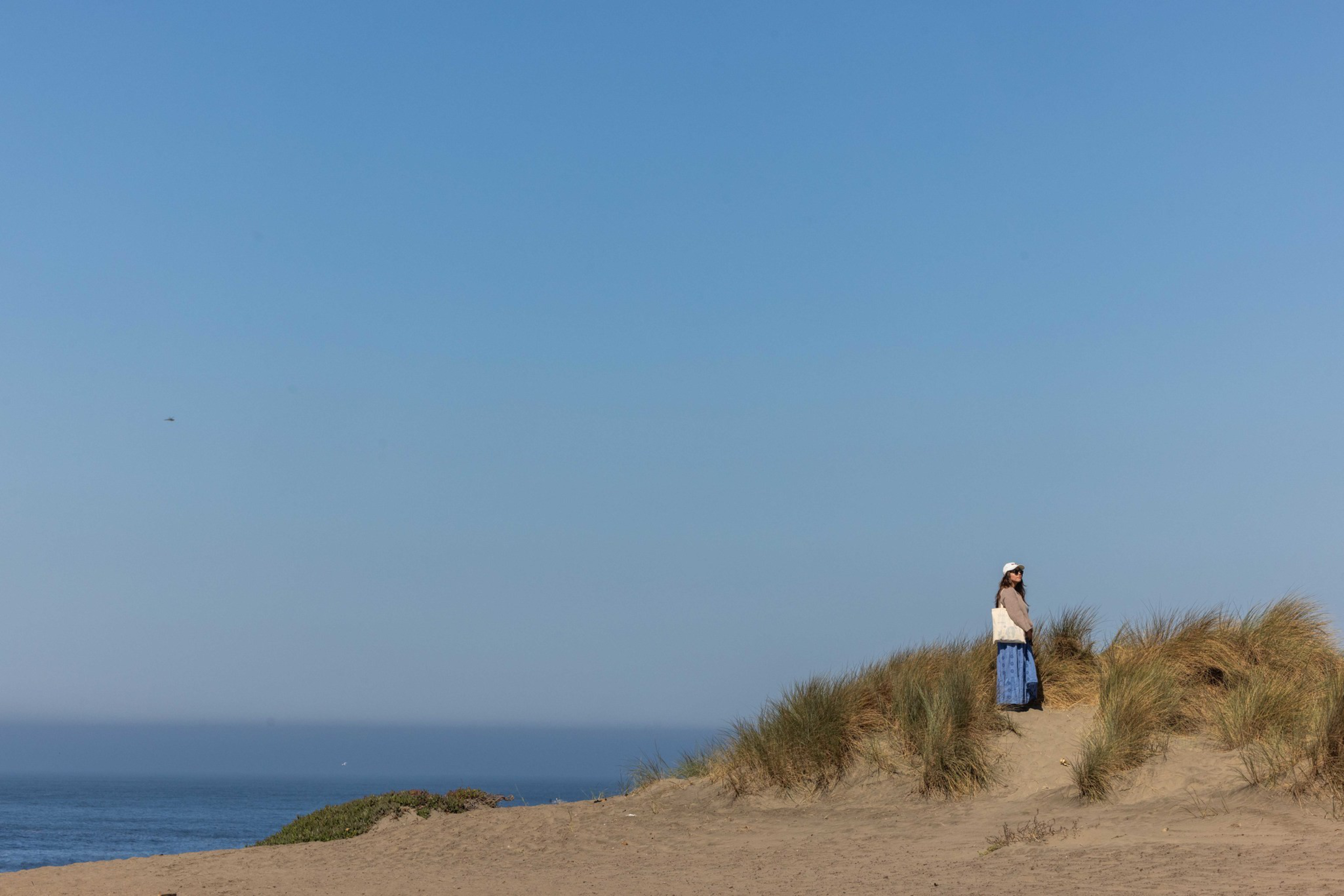 A person wearing a hat and blue skirt stands among sand dunes with dry grass near a calm ocean under a clear blue sky.