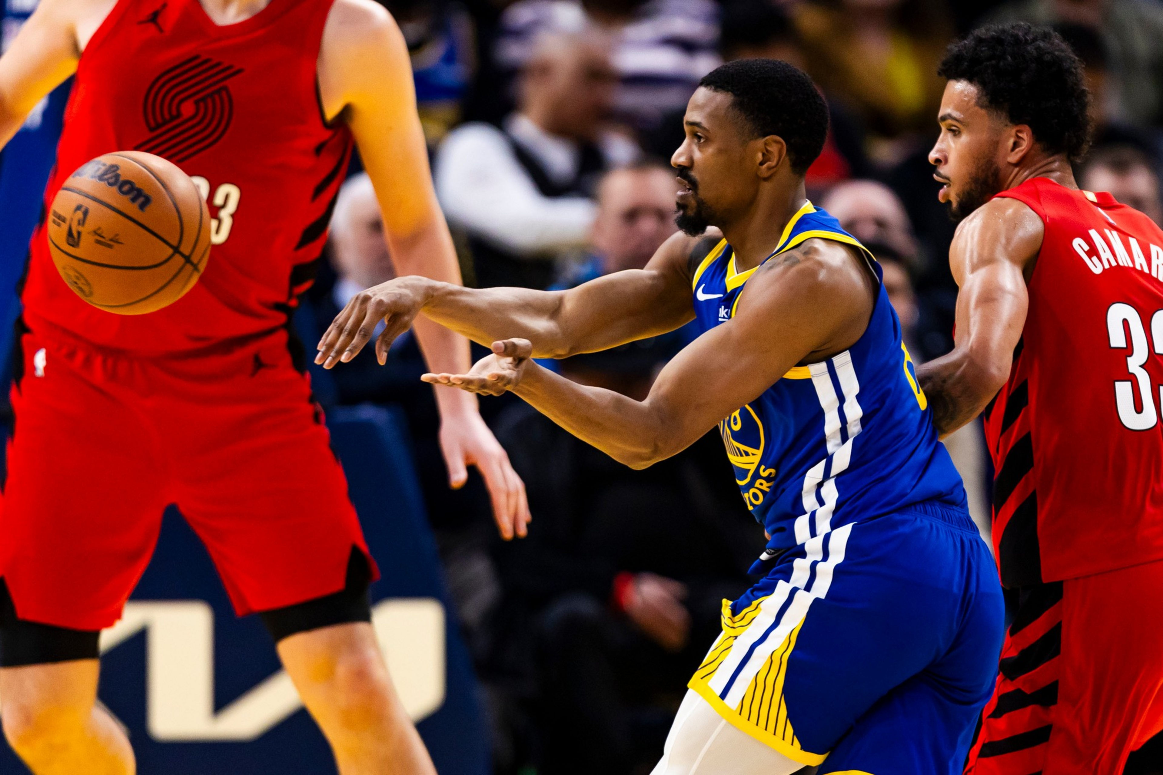 A basketball player in a blue and yellow uniform passes the ball while two players in red uniforms defend nearby.