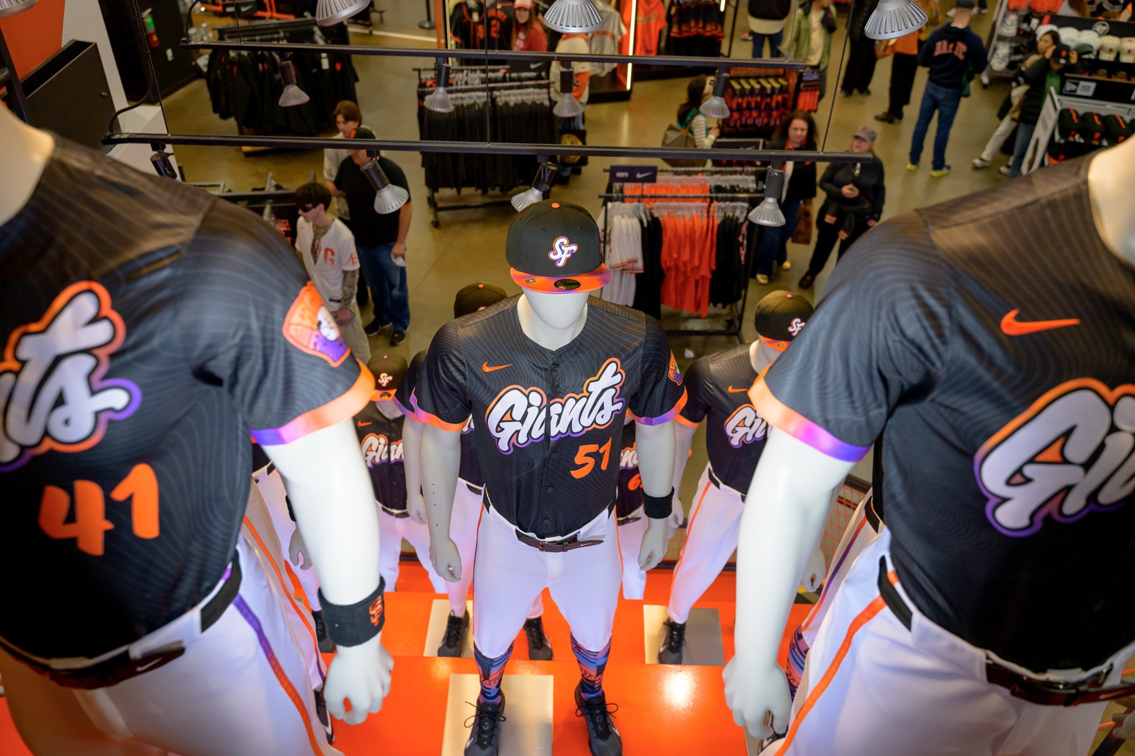 Mannequins wearing black "Giants" baseball jerseys with orange accents are displayed in a store. People shop in the background.