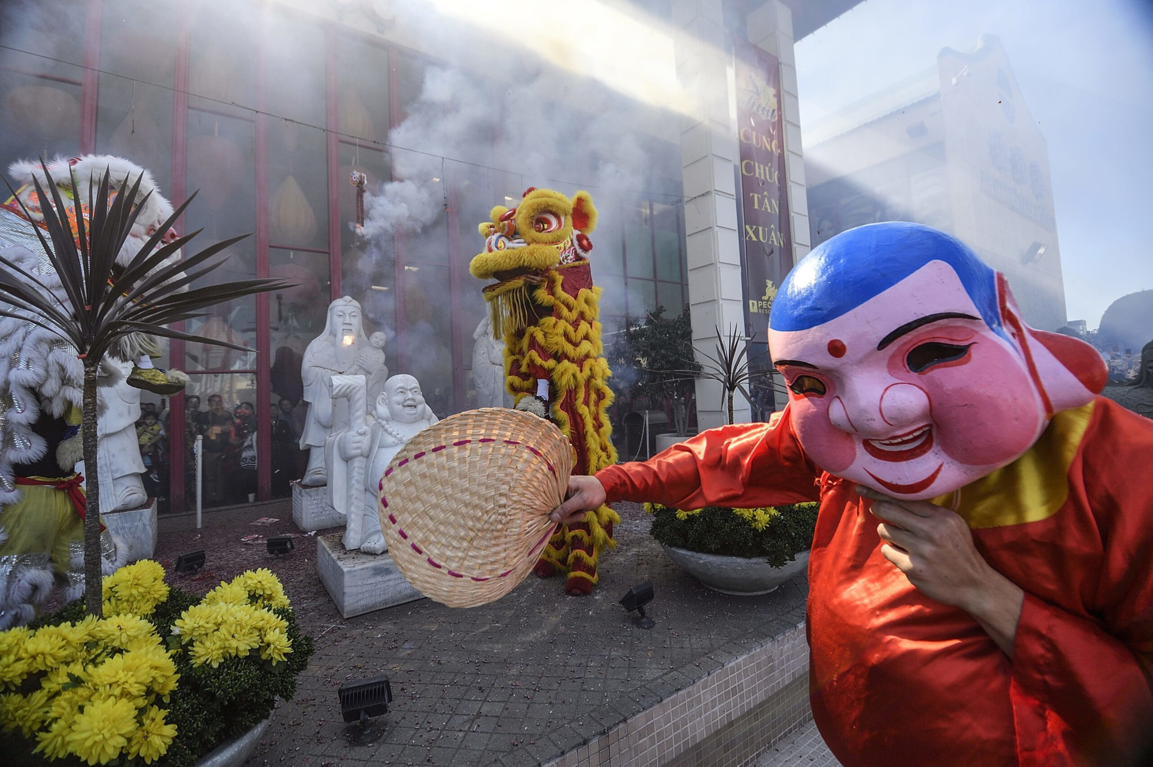 A person wearing a colorful mask and red costume waves a woven fan beside two lion dance performers and stone statues amid smoke and yellow flowers.