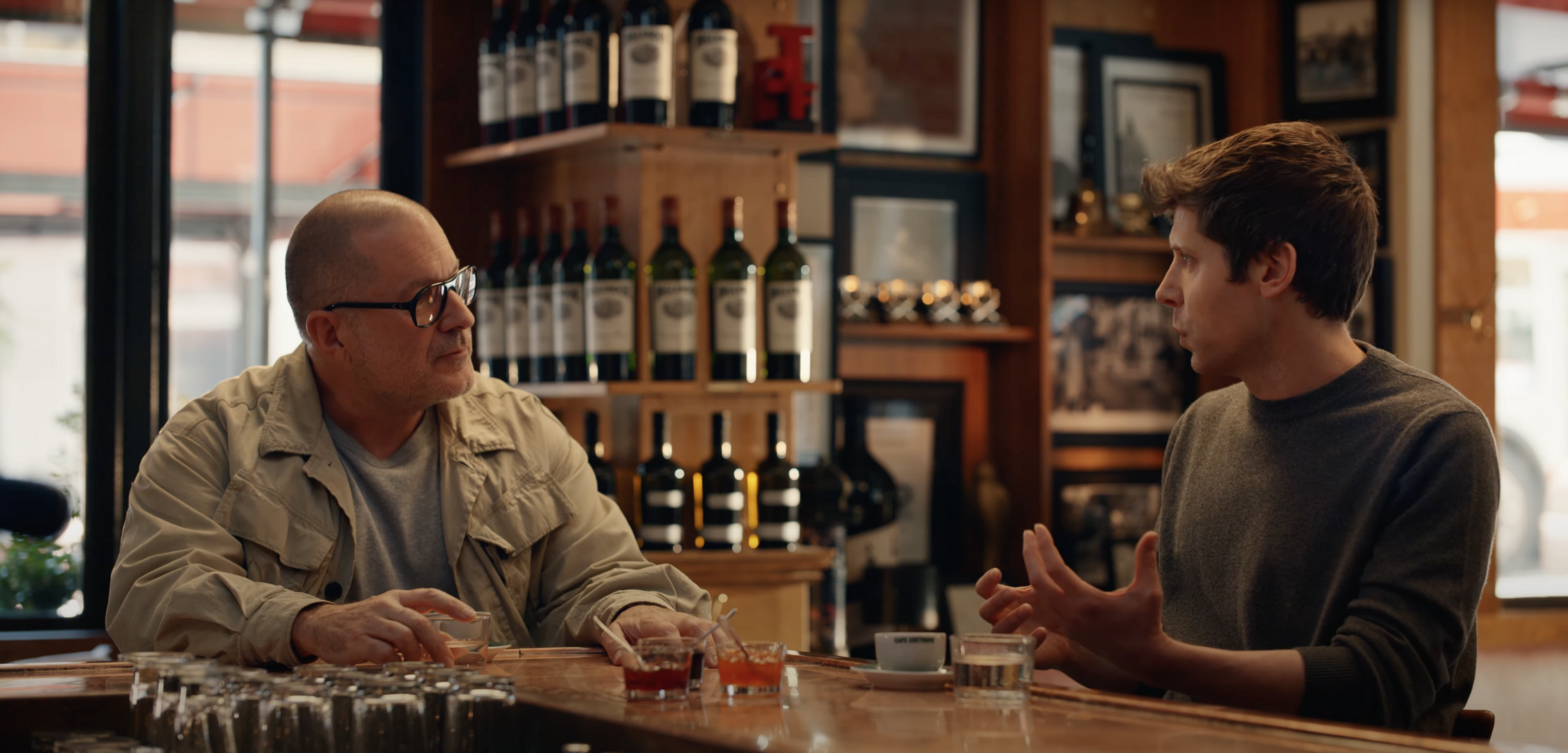 Two men sit at a wooden bar counter, engaged in conversation. Bottles line the shelves behind them, and the setting feels casual and warm.