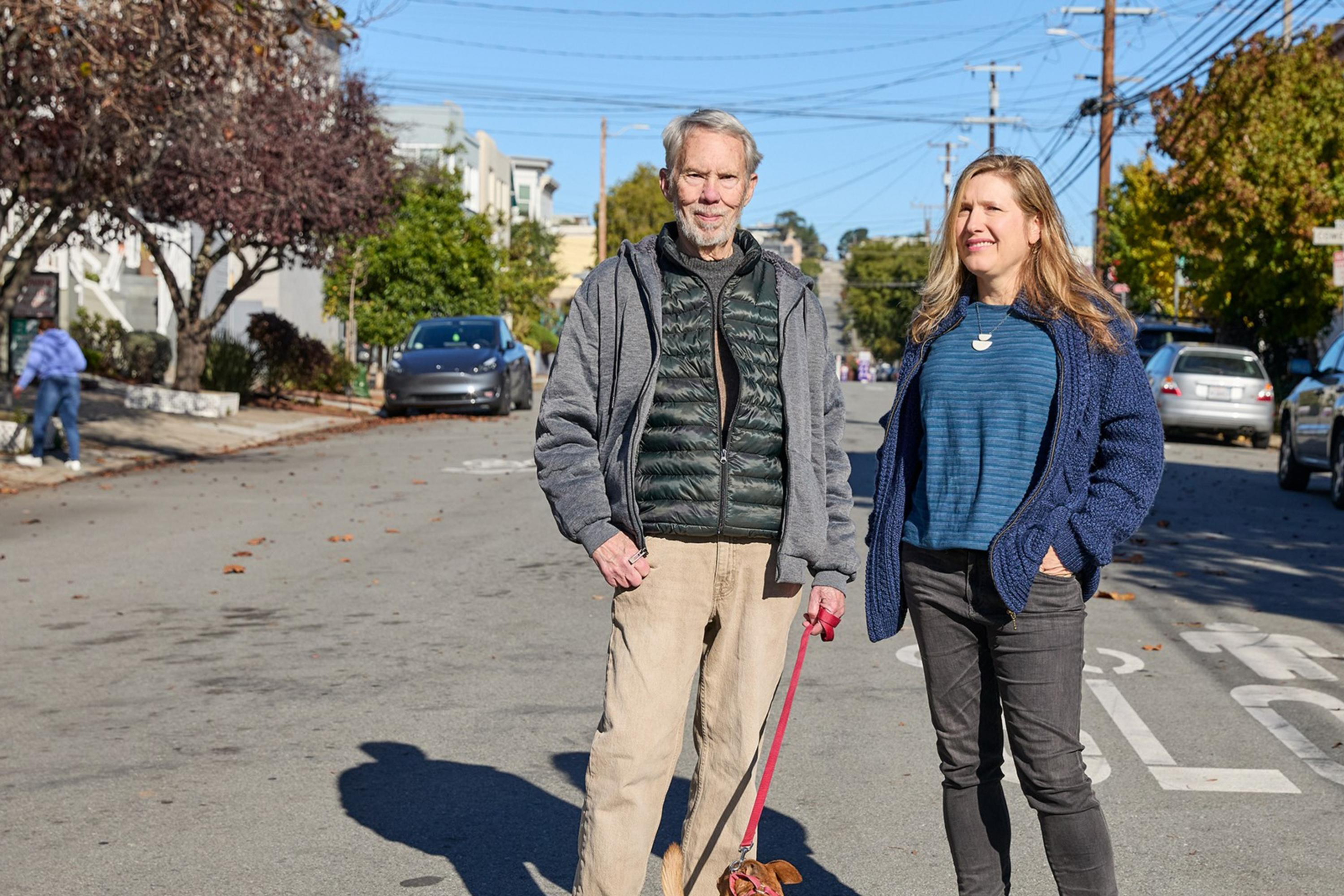 A man and woman stand on a sunlit street with a small dog on a leash. They are casually dressed, with trees and parked cars lining both sides of the street.