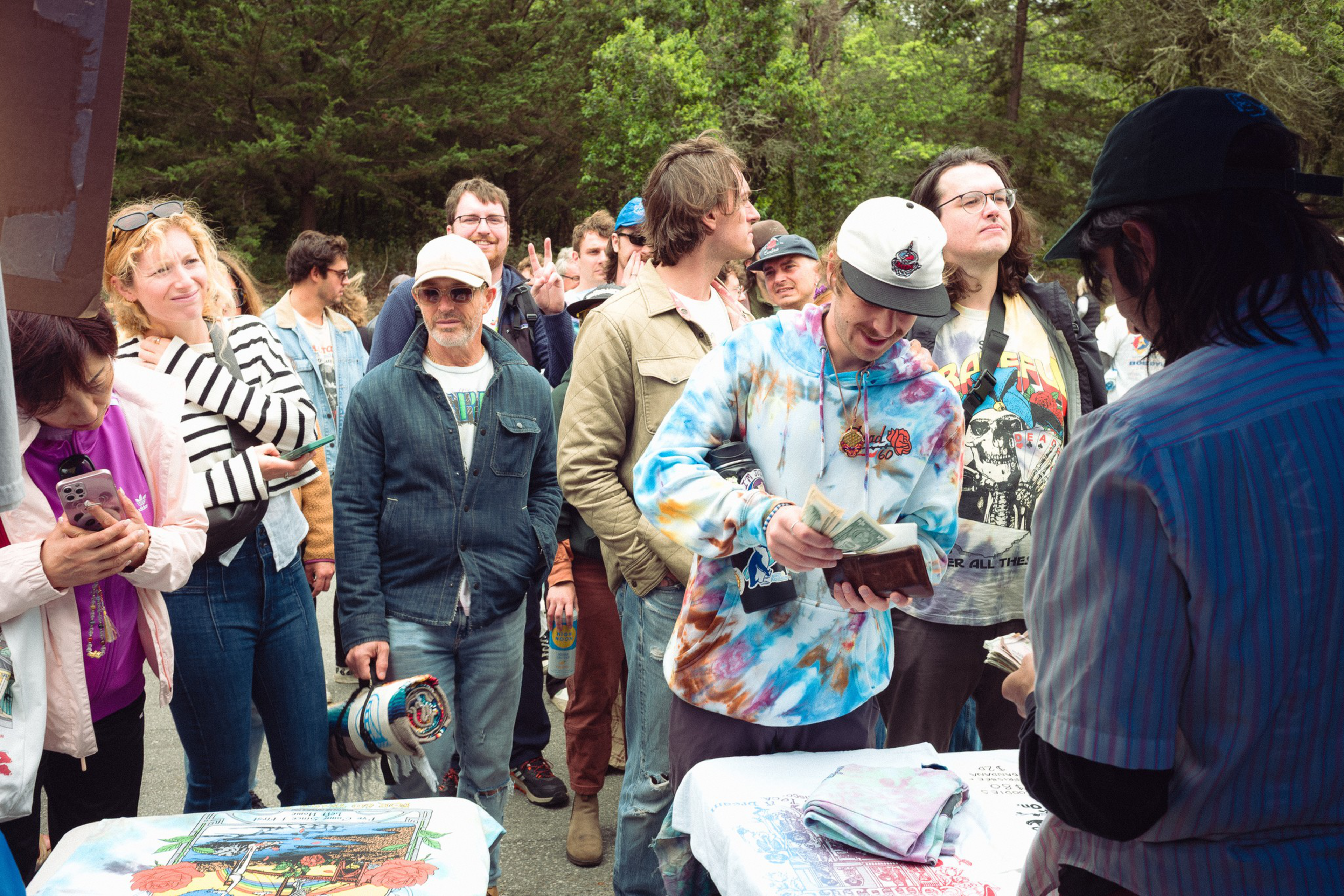 A diverse group of people stands outdoors near tables with colorful shirts, one person in a tie-dye hoodie counts money while others watch or use their phones.