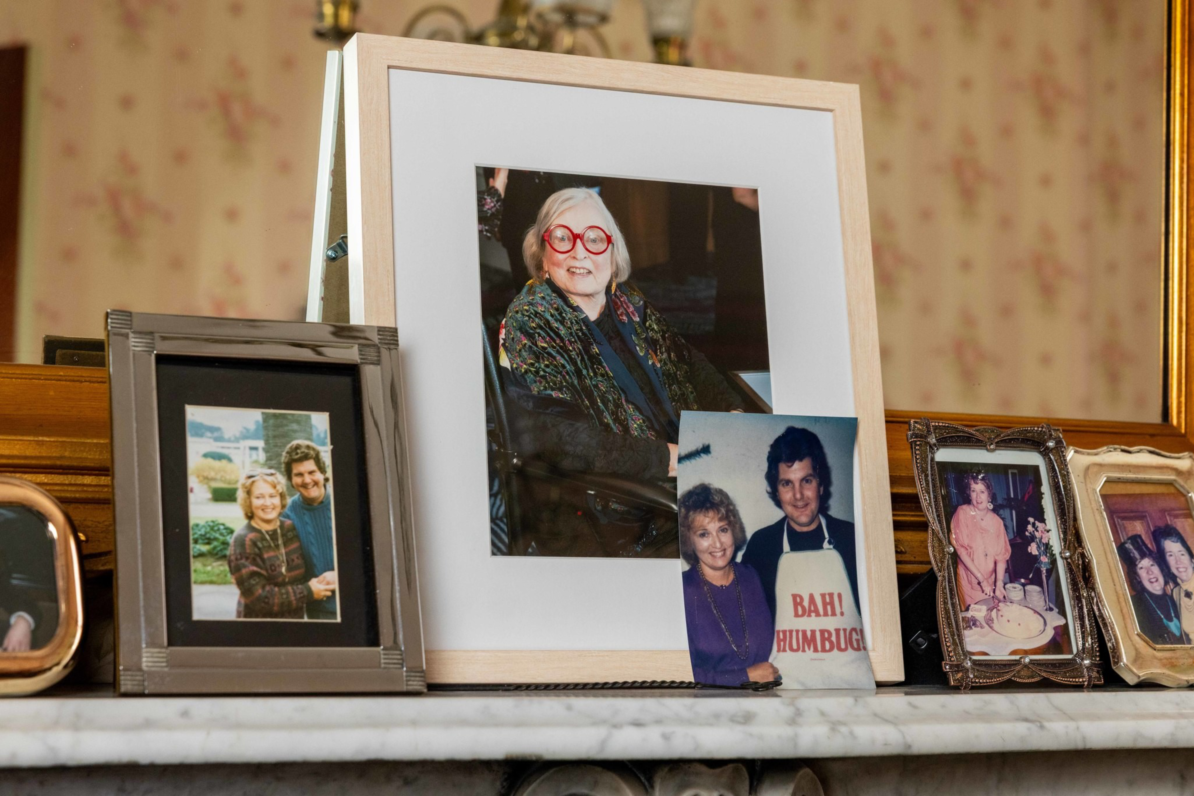 Several framed family photos rest on a mantle, including a woman with red glasses, a couple in front of a house, and a man wearing a “BAH! HUMBUG” apron.