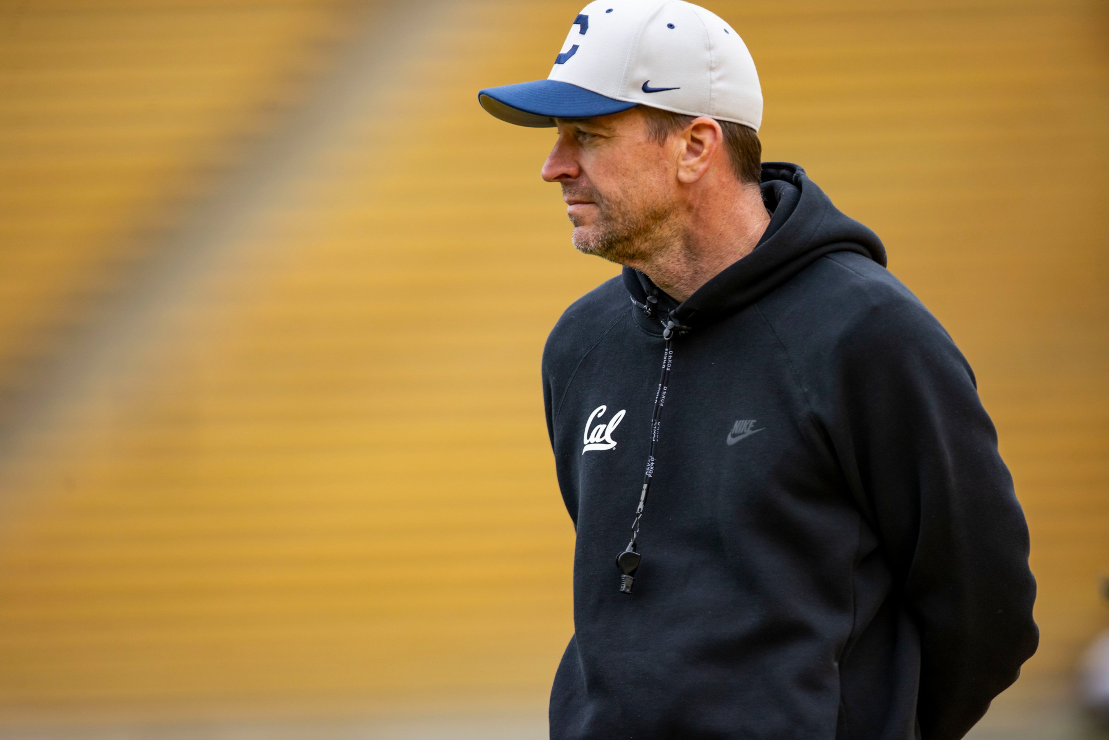 A man wearing a white and blue cap, black Cal hoodie with a whistle around his neck, stands on a field with yellow bleachers in the background.
