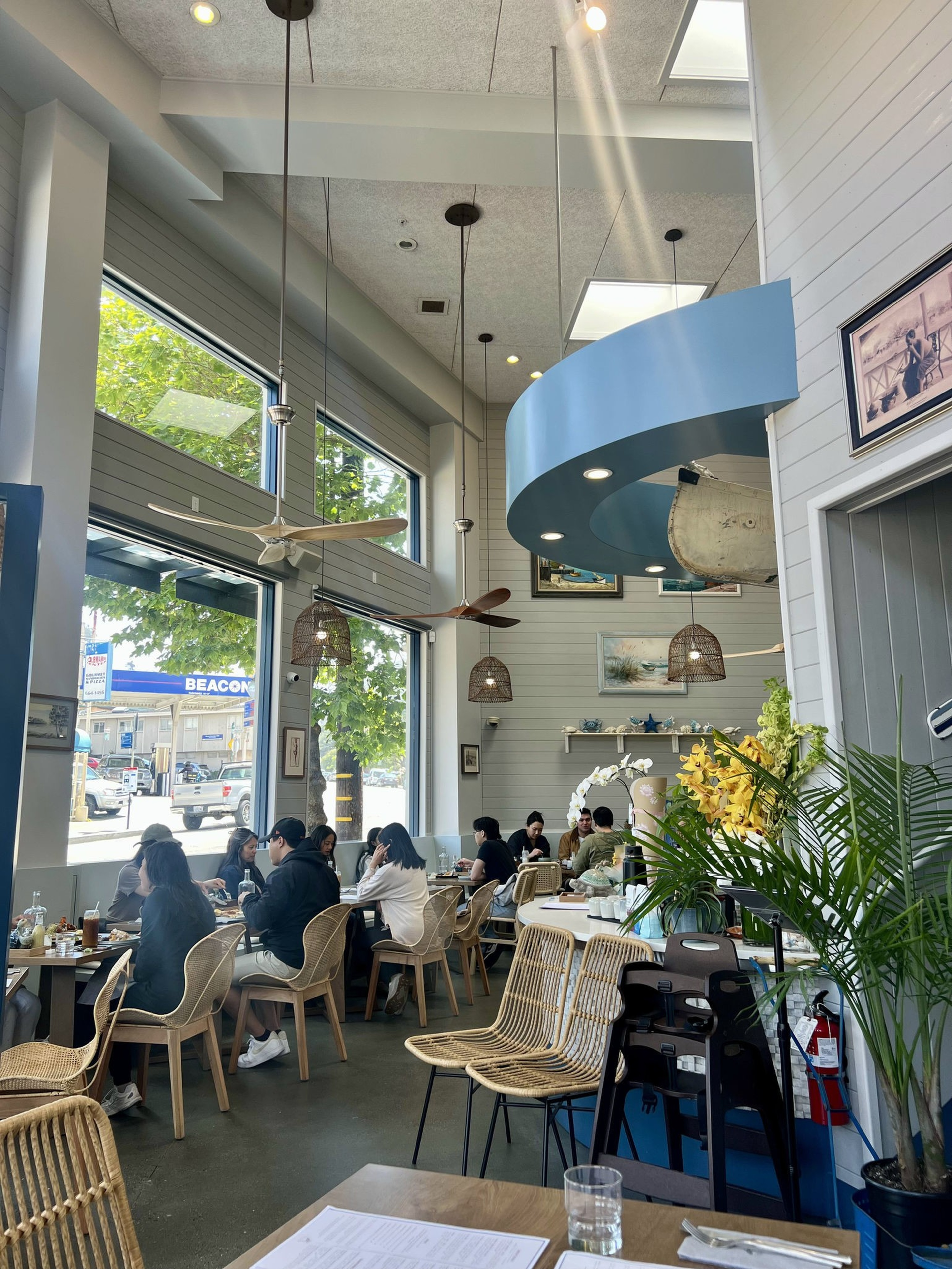 Customers sit and chat at a cozy, well-lit cafe with wicker chairs, large windows, hanging fans, and blue ceiling accents.