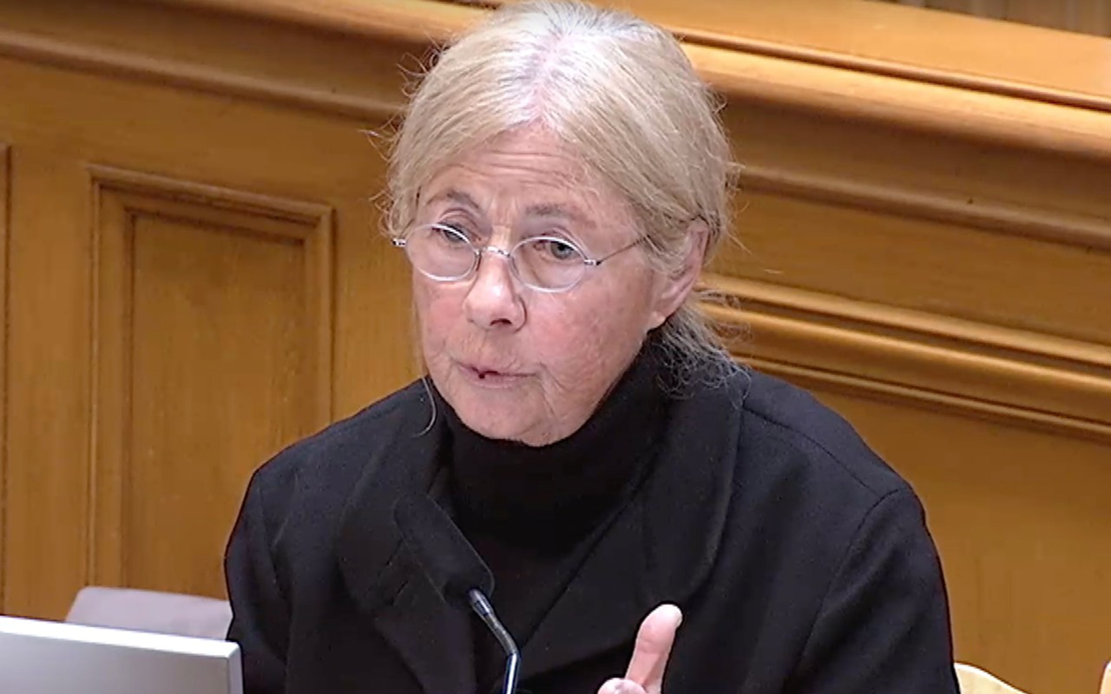 A person with light hair and glasses is speaking, seated indoors. They are wearing a black jacket and are positioned in front of a laptop next to a wooden wall.