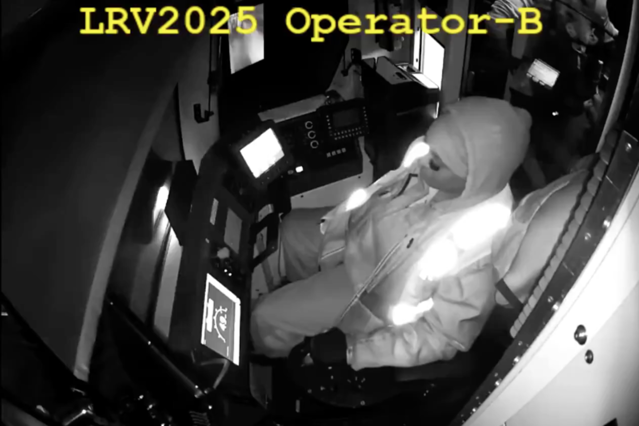 A person dressed in a hooded jacket and gloves sits in a control cabin, monitoring illuminated screens, with another person holding a tablet nearby.