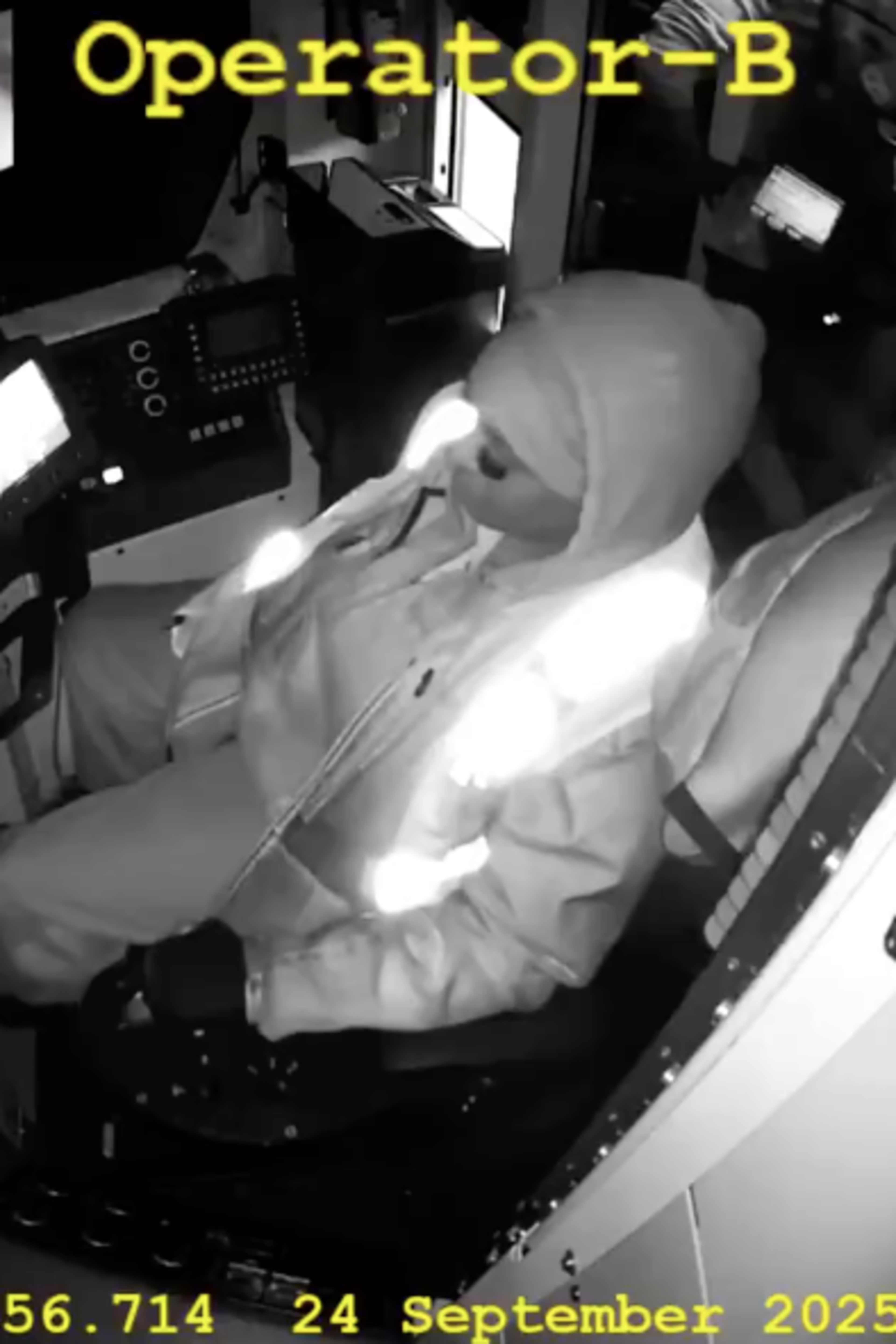 A person dressed in a hooded jacket and gloves sits in a control cabin, monitoring illuminated screens, with another person holding a tablet nearby.