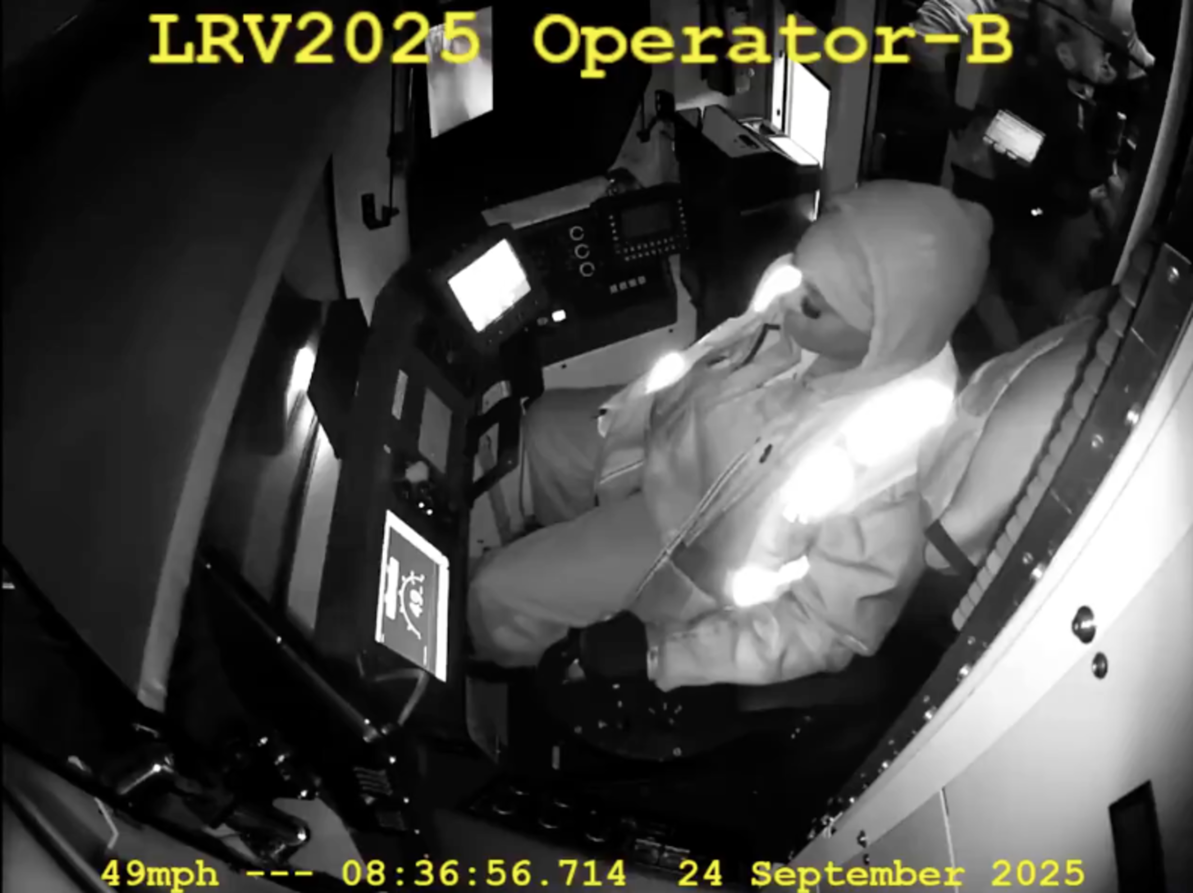 A person dressed in a hooded jacket and gloves sits in a control cabin, monitoring illuminated screens, with another person holding a tablet nearby.
