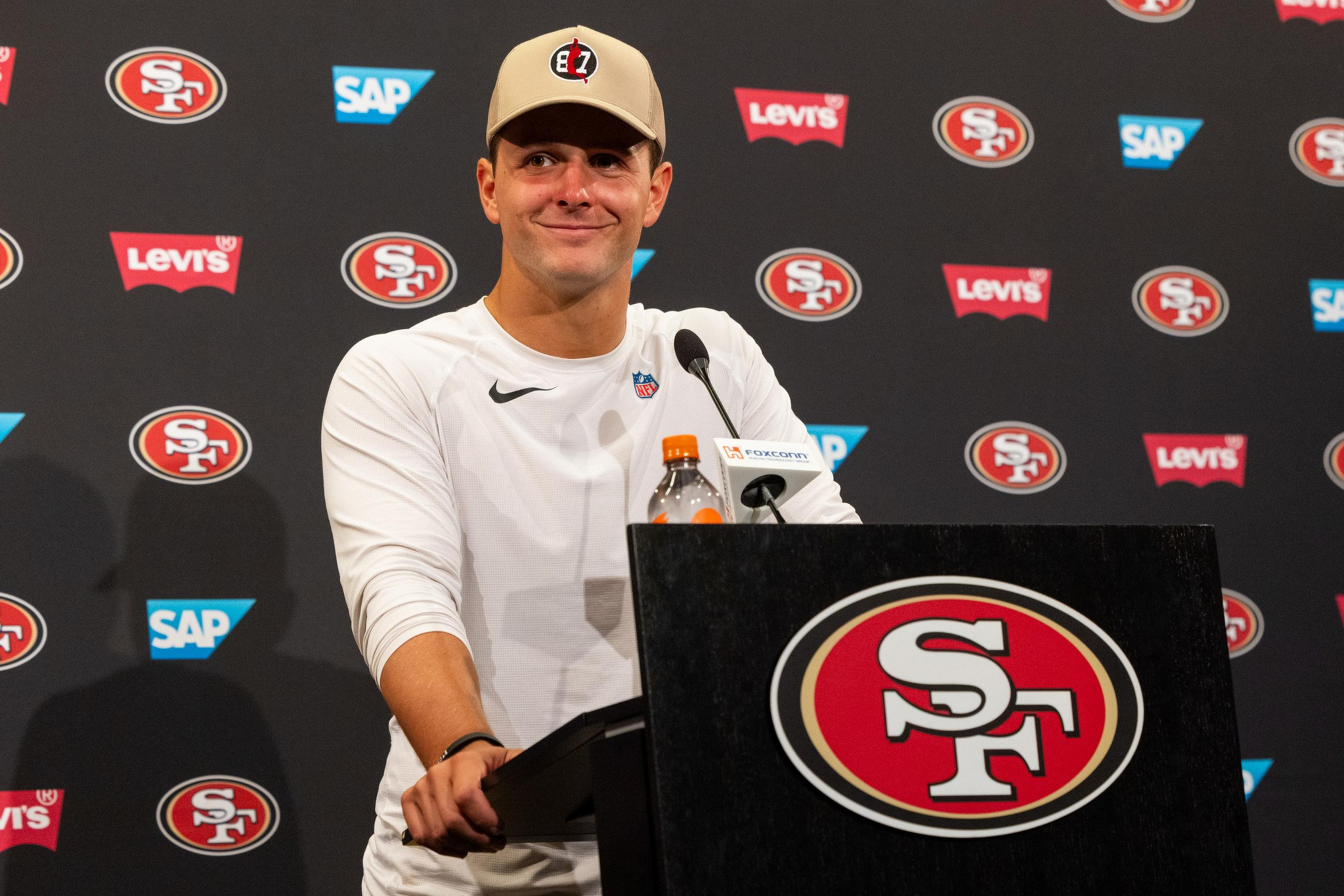 A man in a white Nike shirt and beige cap stands at a podium with San Francisco 49ers logos and sponsor logos in the background.