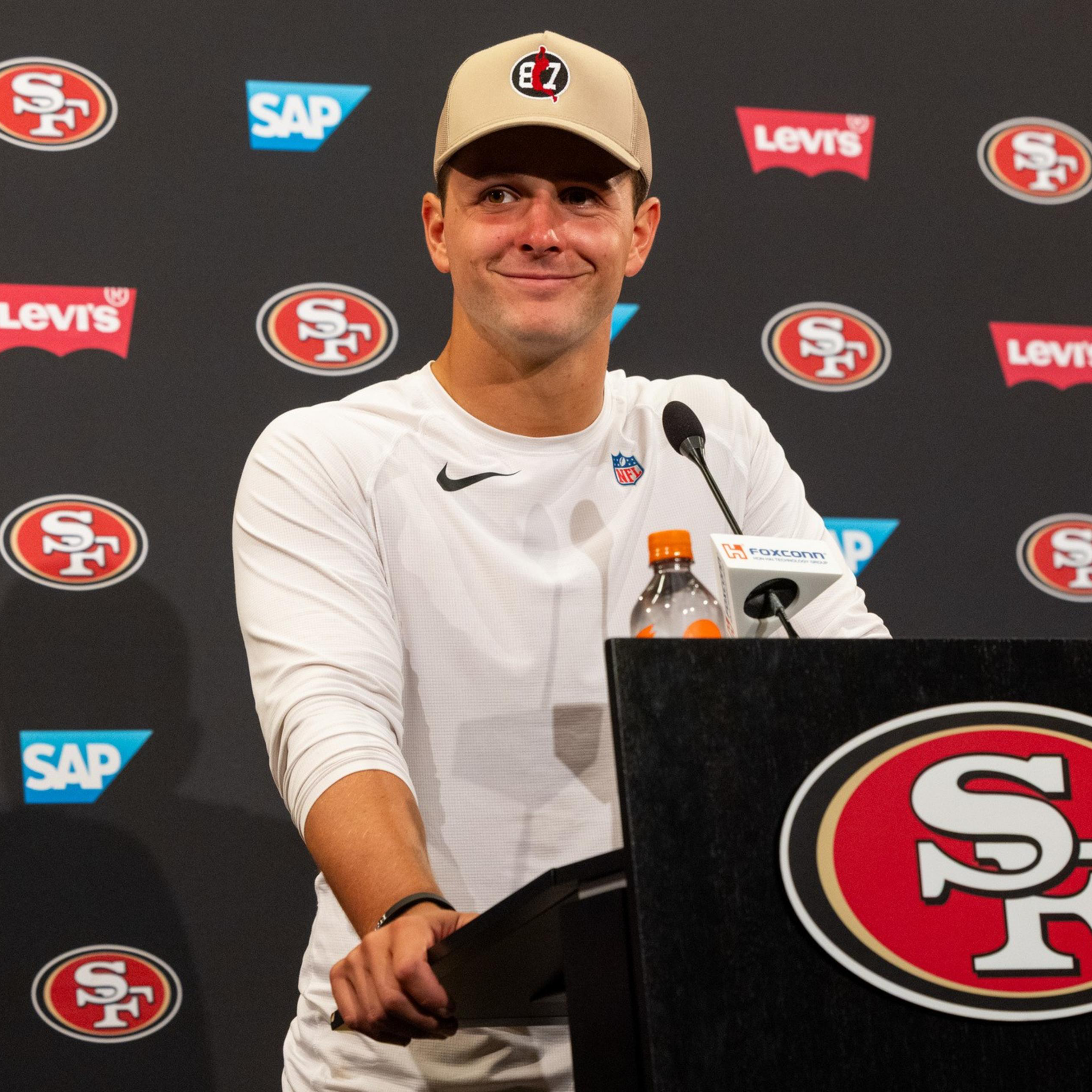 A man in a white Nike shirt and beige cap stands at a podium with San Francisco 49ers logos and sponsor logos in the background.