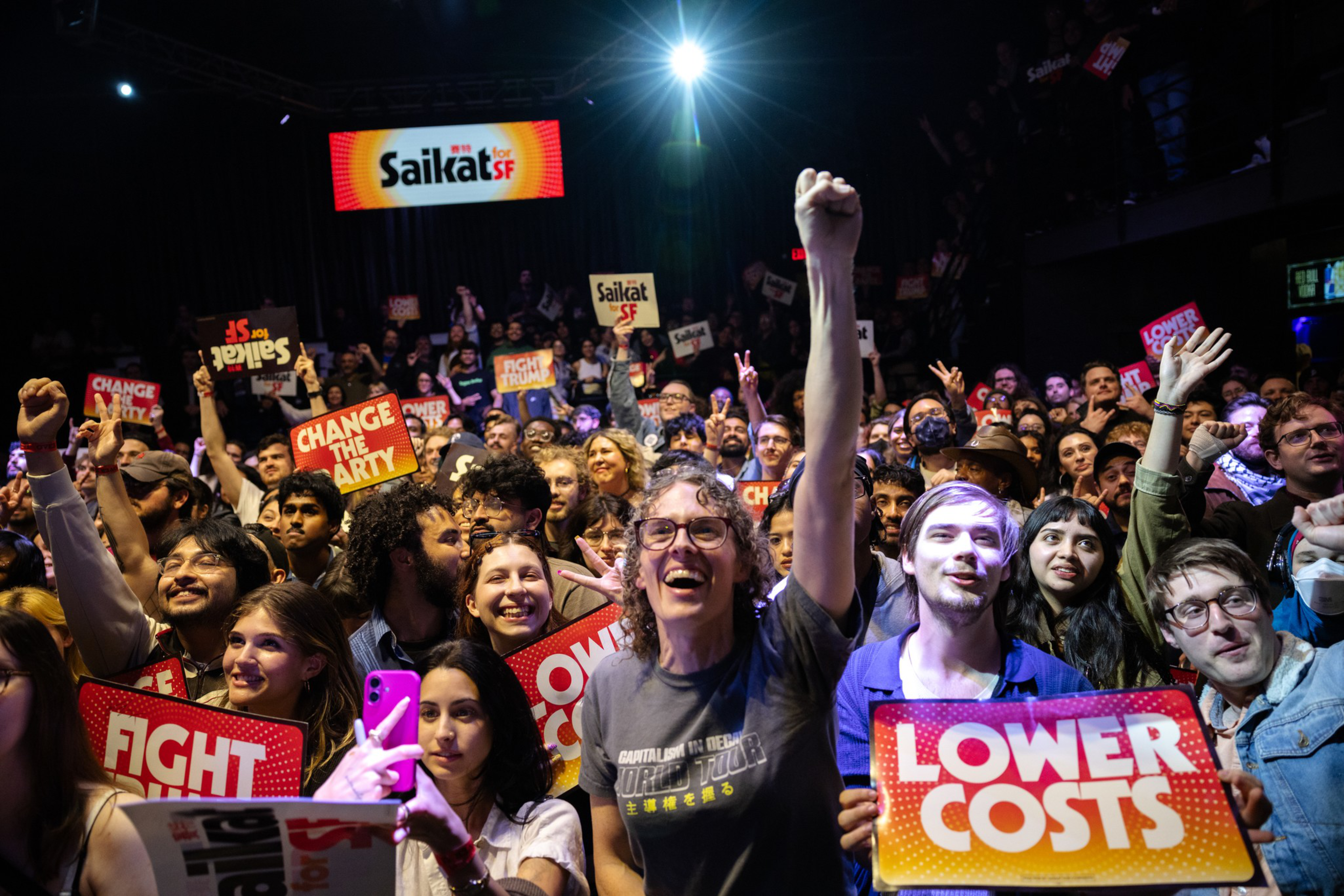 A large, diverse crowd holds up signs with slogans like “FIGHT,” “CHANGE THE PARTY,” and “LOWER COSTS,” cheering enthusiastically at a Saikat SF event.