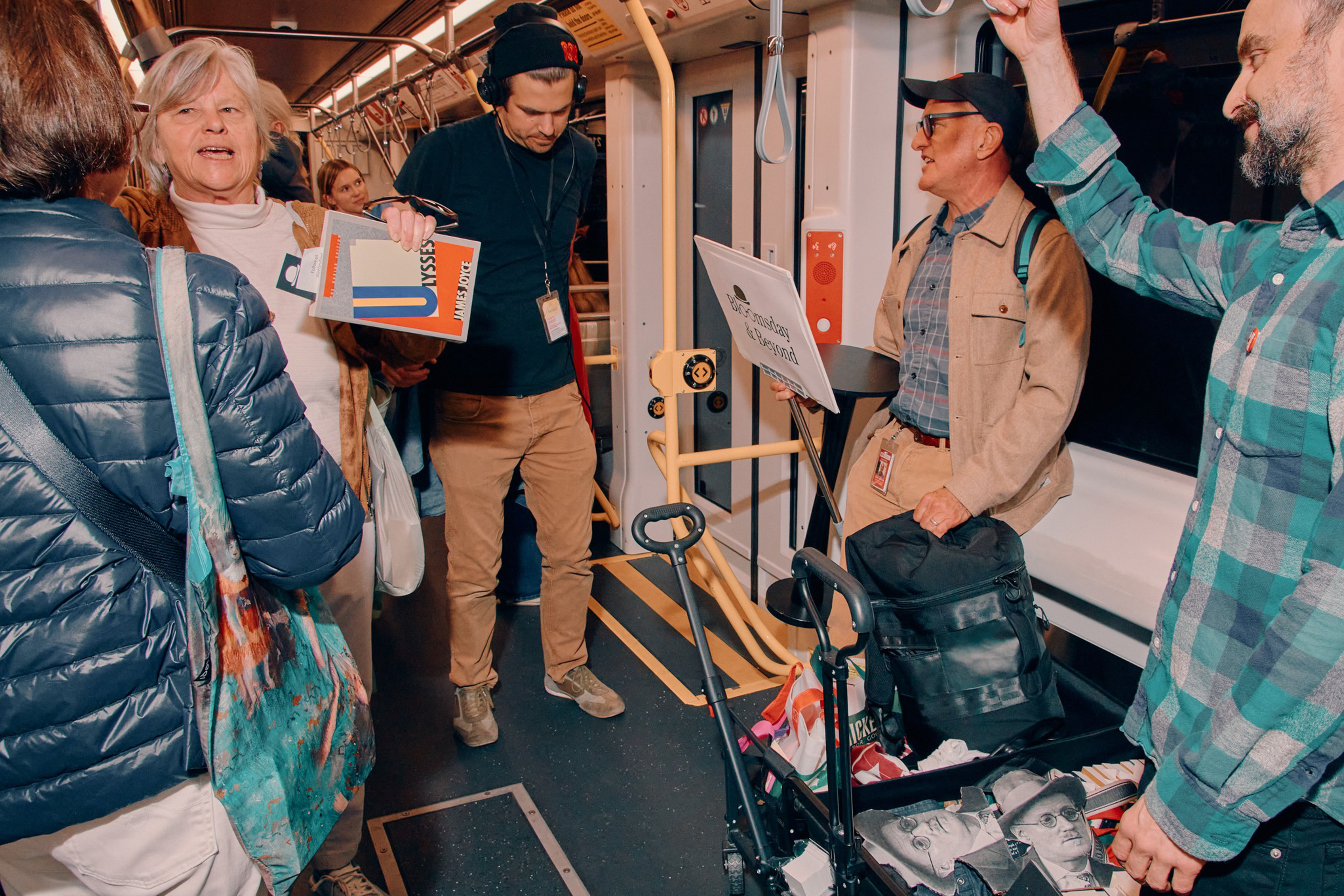 Five people stand inside a subway car, one holding sheet music, another with headphones, and a cart with books and a portrait sketch.