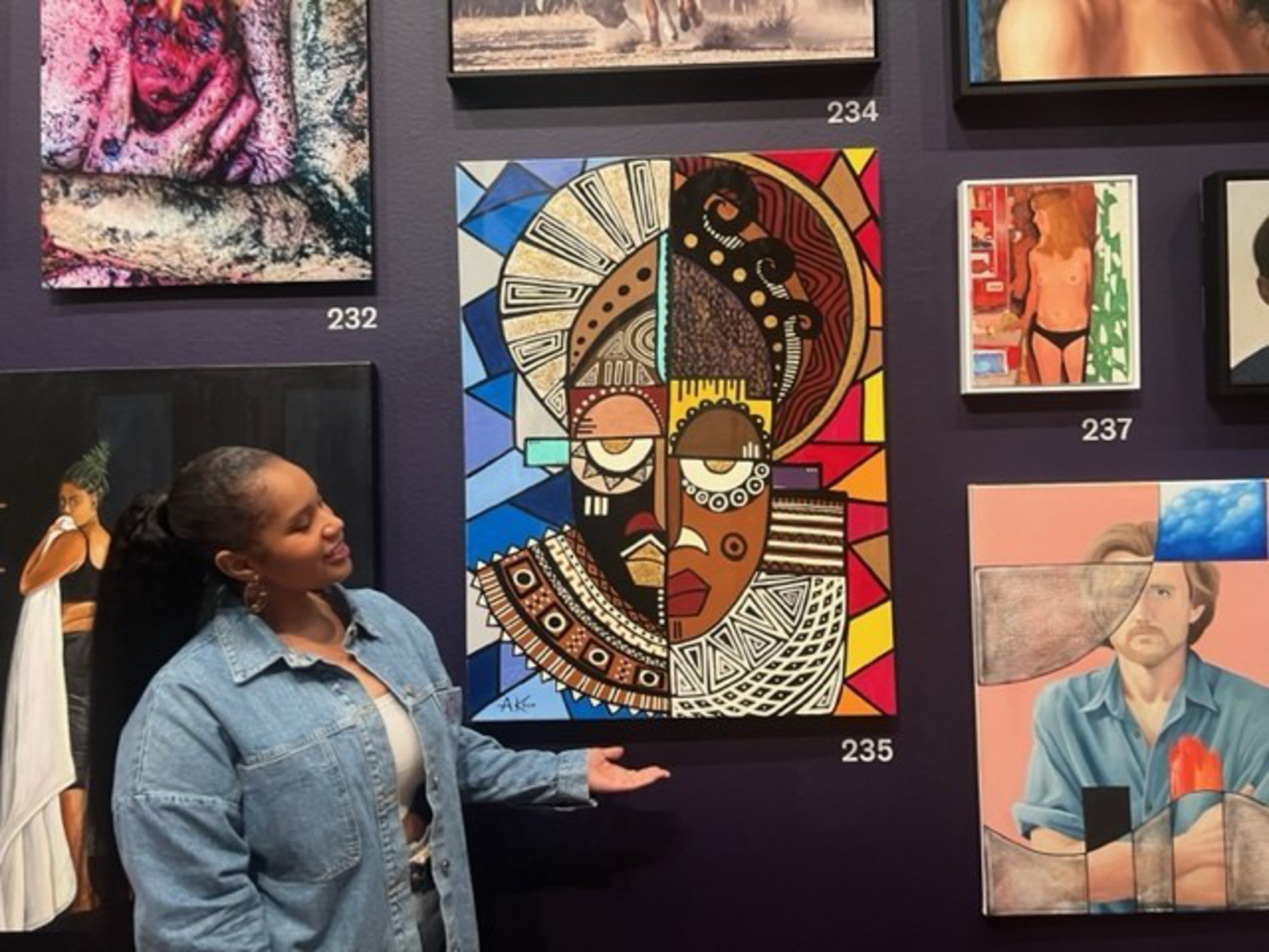 A woman gestures toward a colorful, abstract painting combining two stylized faces with geometric patterns, surrounded by other varied artworks on a dark wall.