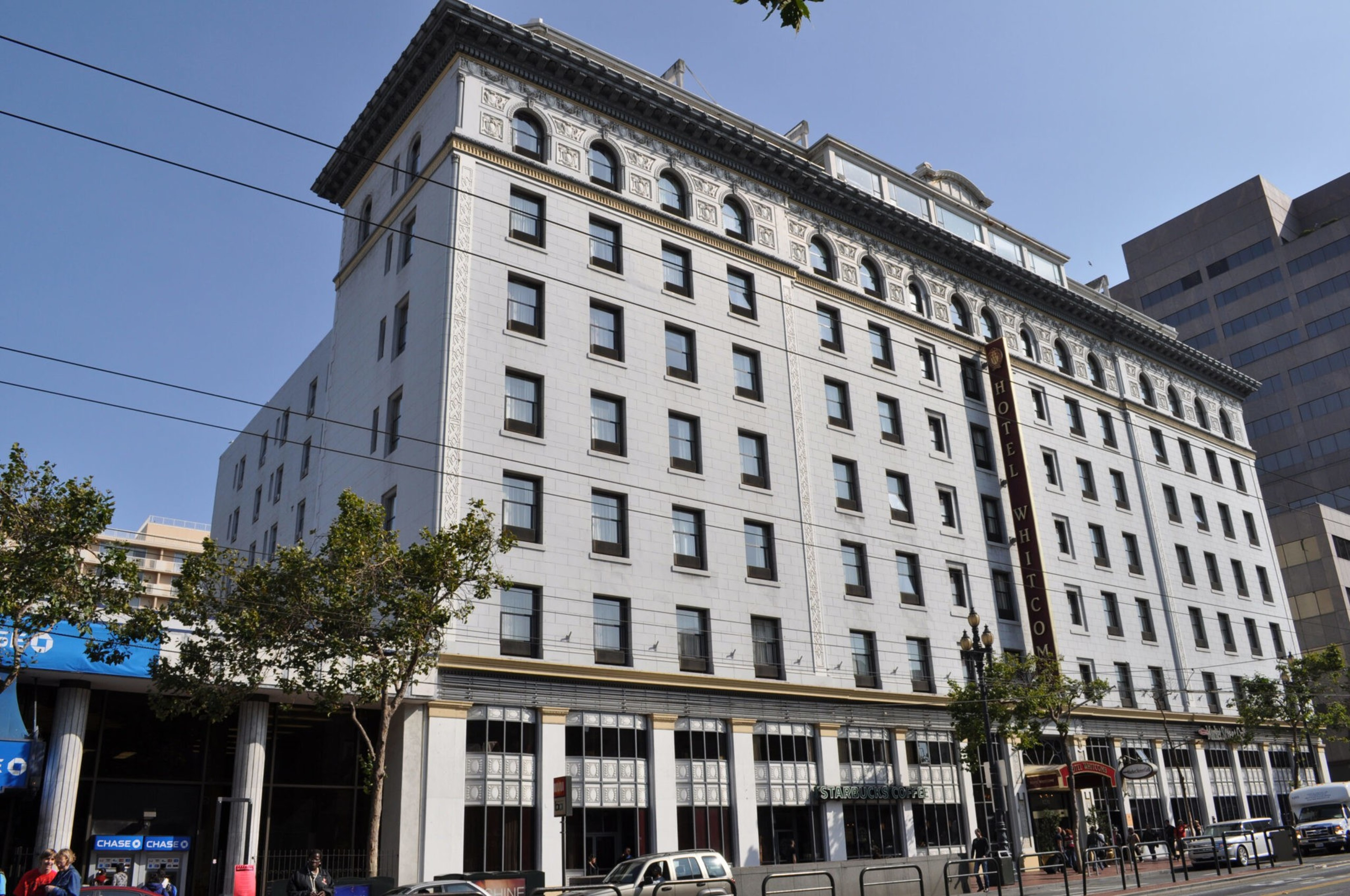 A large, white, rectangular hotel building with multiple windows, ornate upper trim, trees lining the sidewalk, and a street with parked cars.