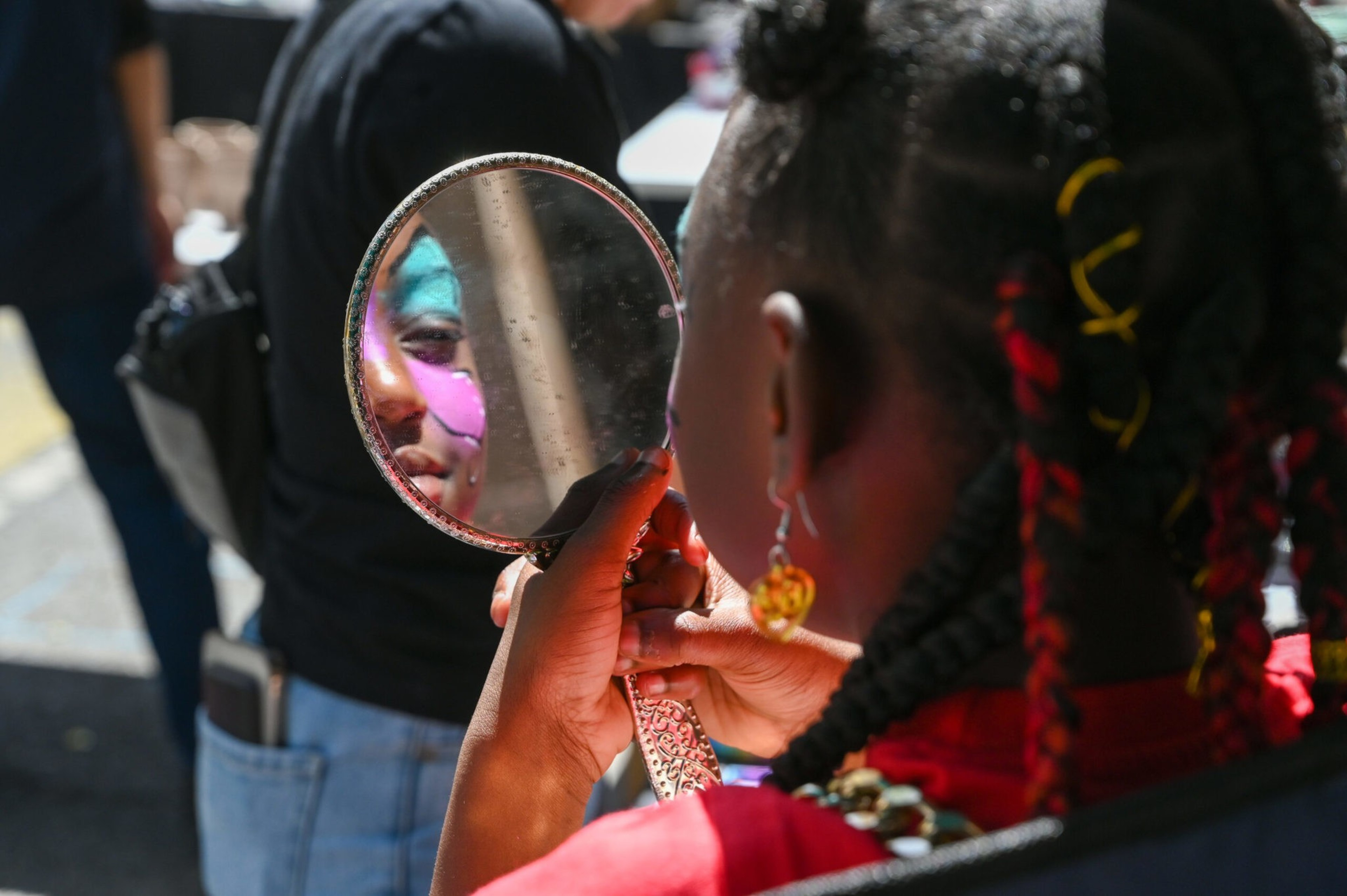 A person with braided hair and face paint looks into a handheld mirror, reflecting their colorful makeup.