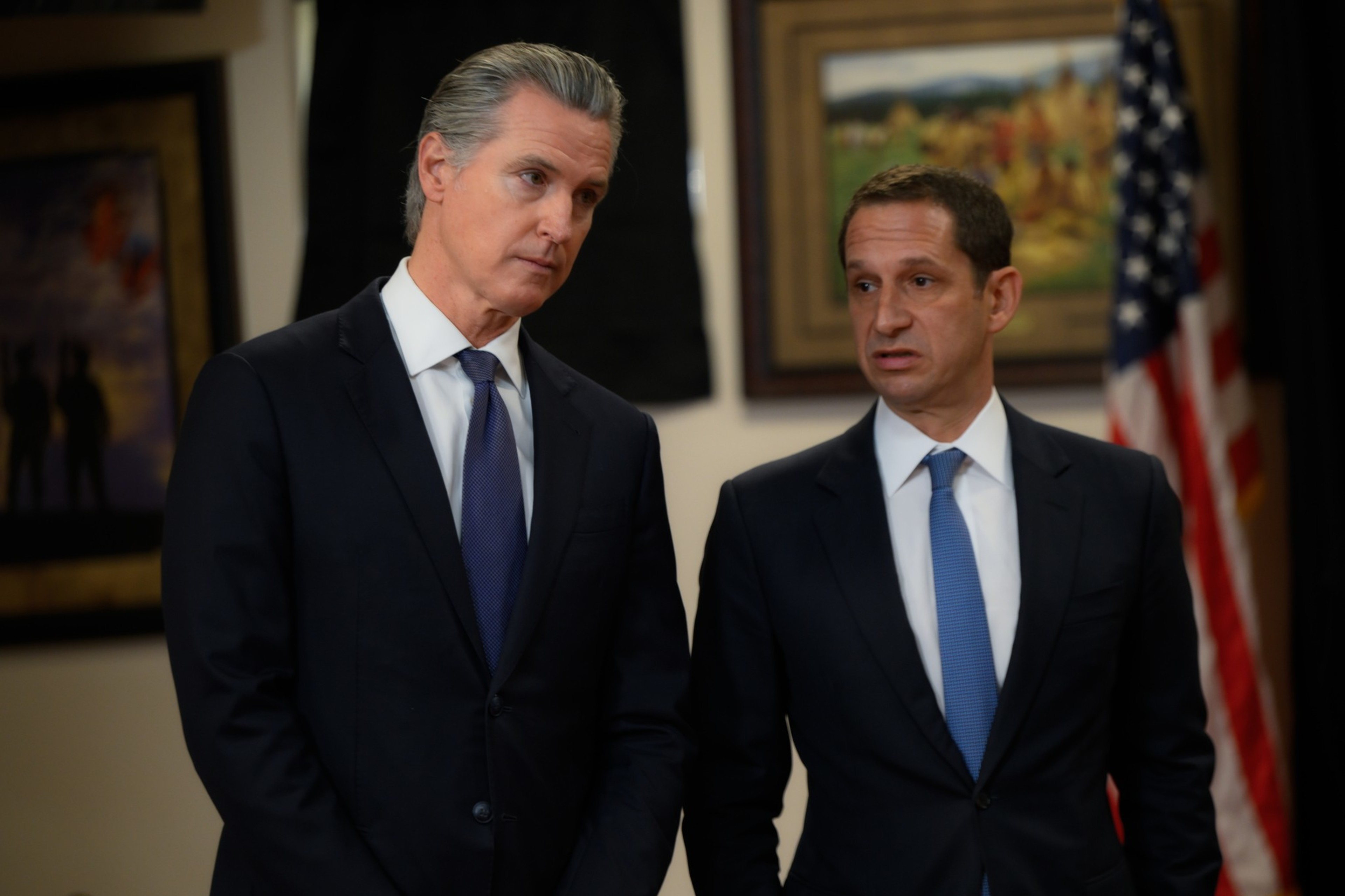 Two men in dark suits and ties stand in an office with framed artwork and a partially visible American flag behind them.