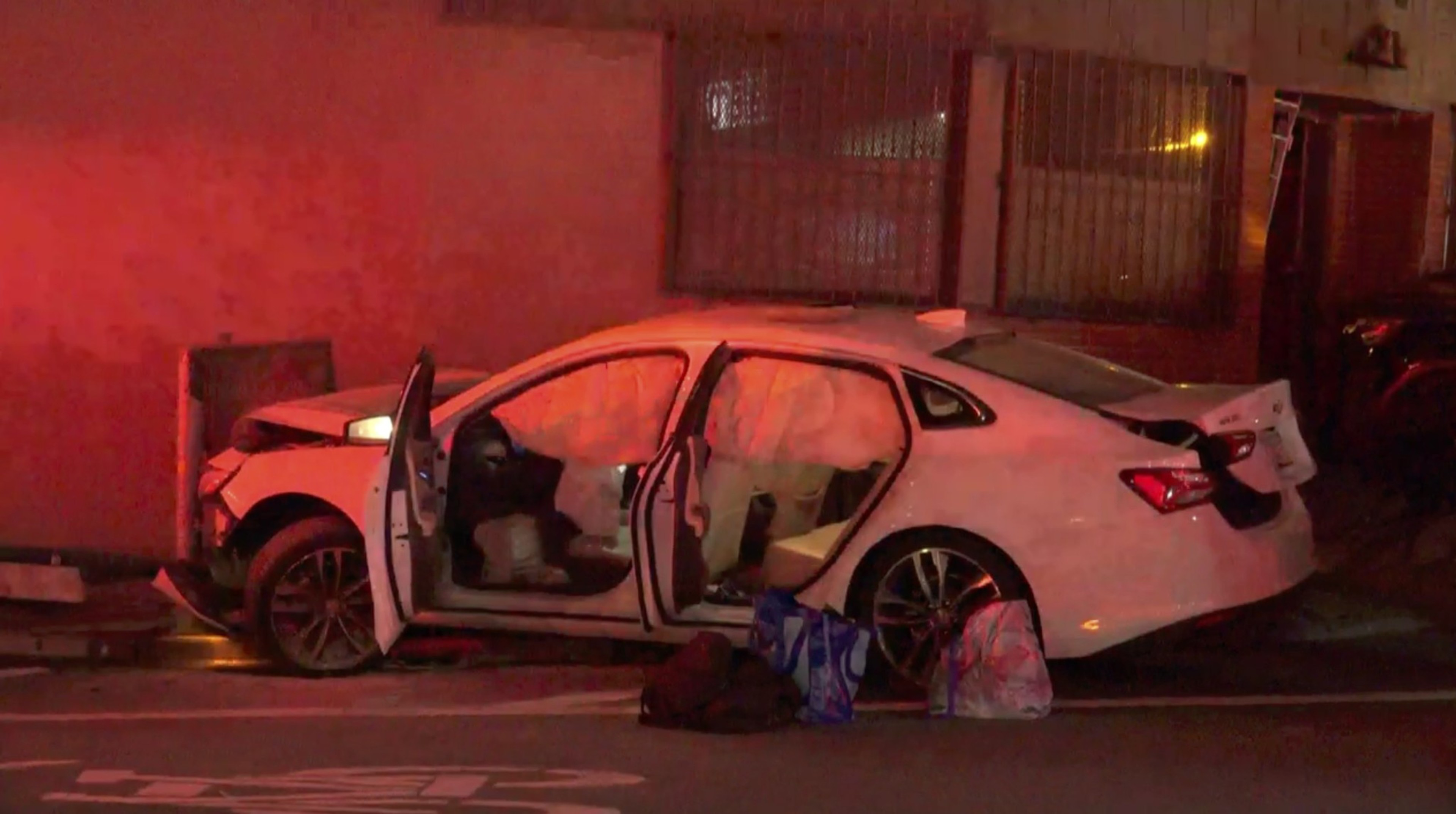 A white car is crashed against a building with airbags deployed and doors open. Several bags are on the ground beside it, and the scene is lit by emergency lights.