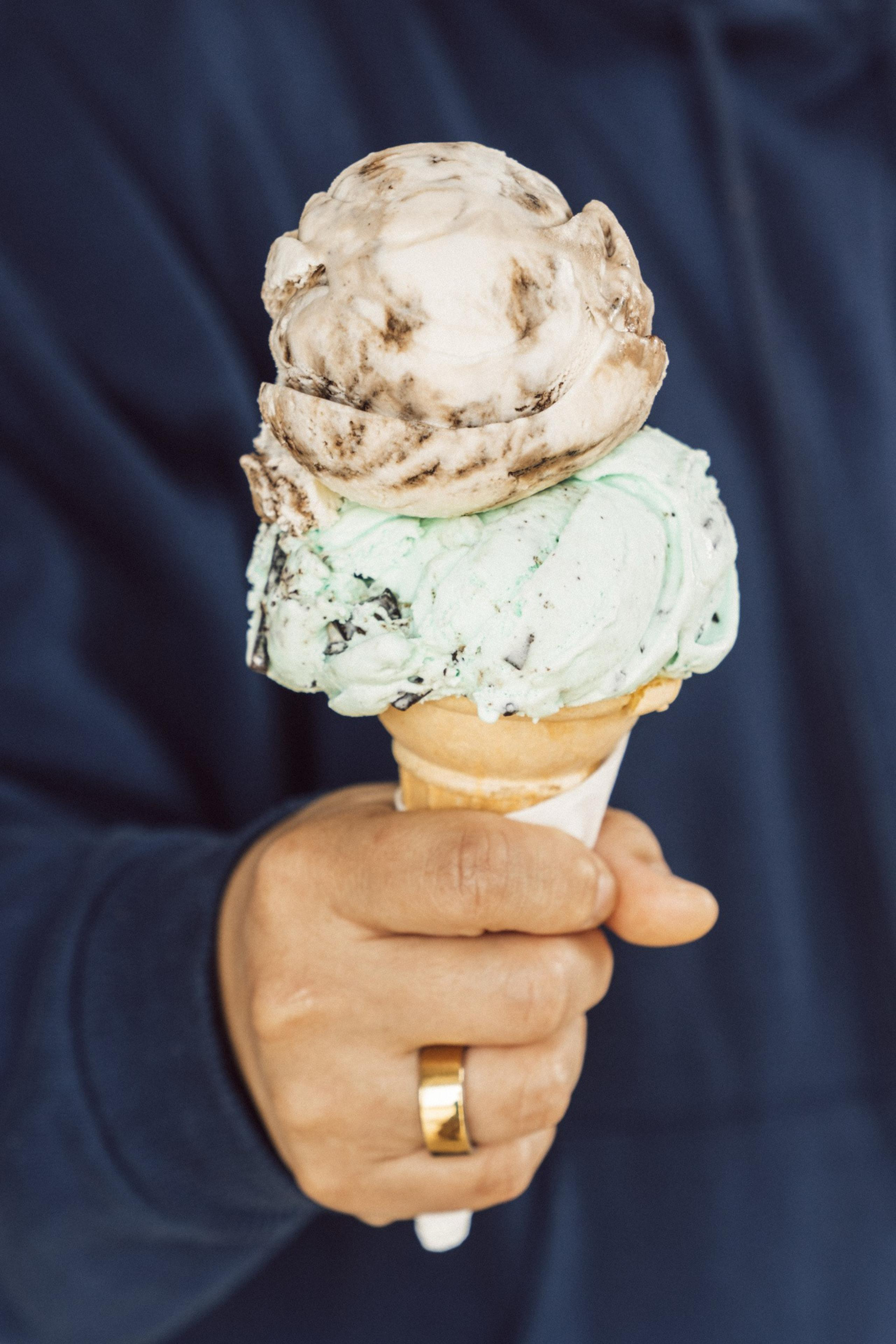A hand holds an ice cream cone with two scoops: mint chocolate chip on the bottom and a swirled brown and white scoop on top. A gold ring is on the finger.