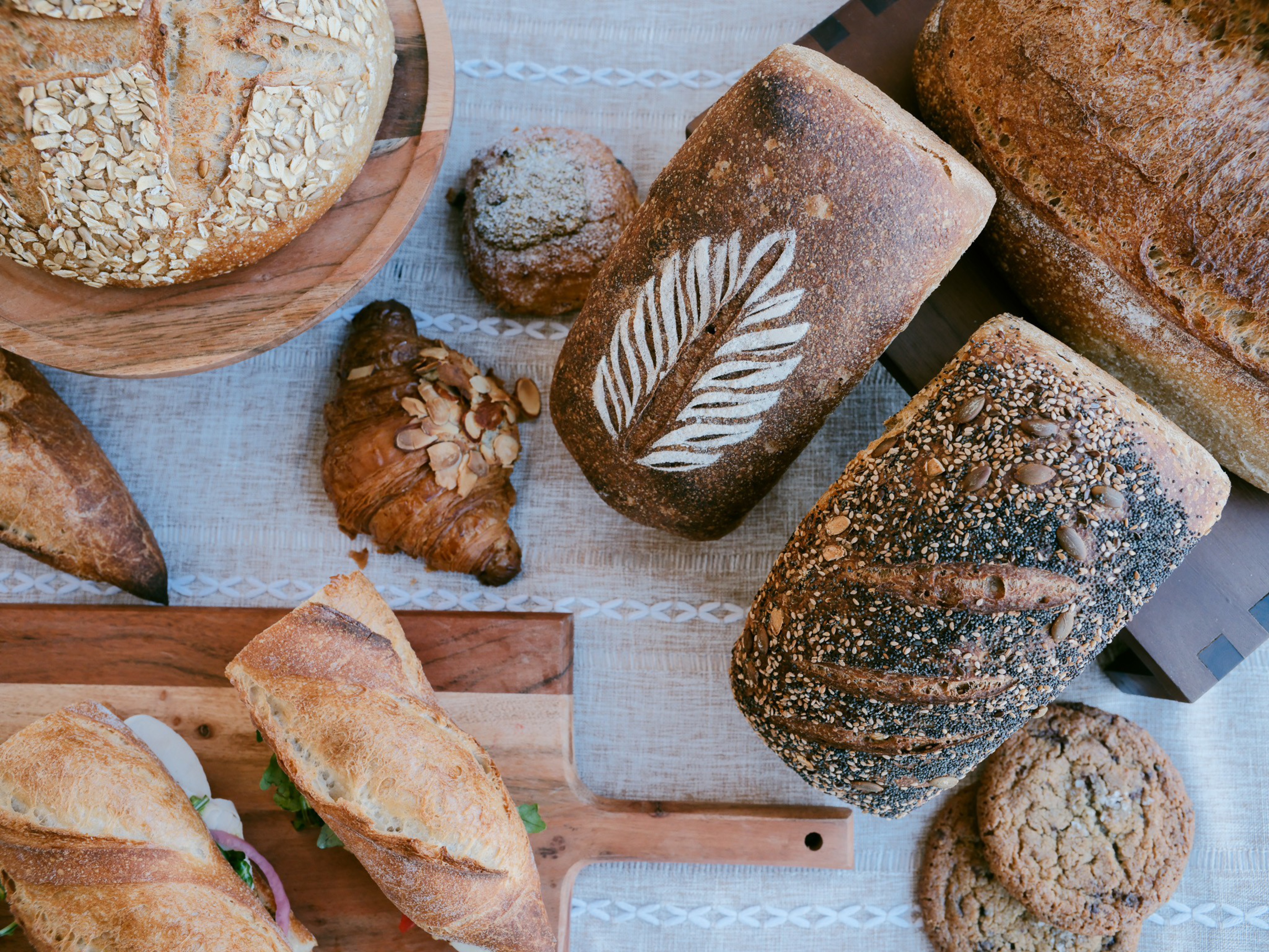 Various types of bread, a croissant topped with almond slices, two sandwich baguettes, and two cookies are arranged on a linen-covered table.