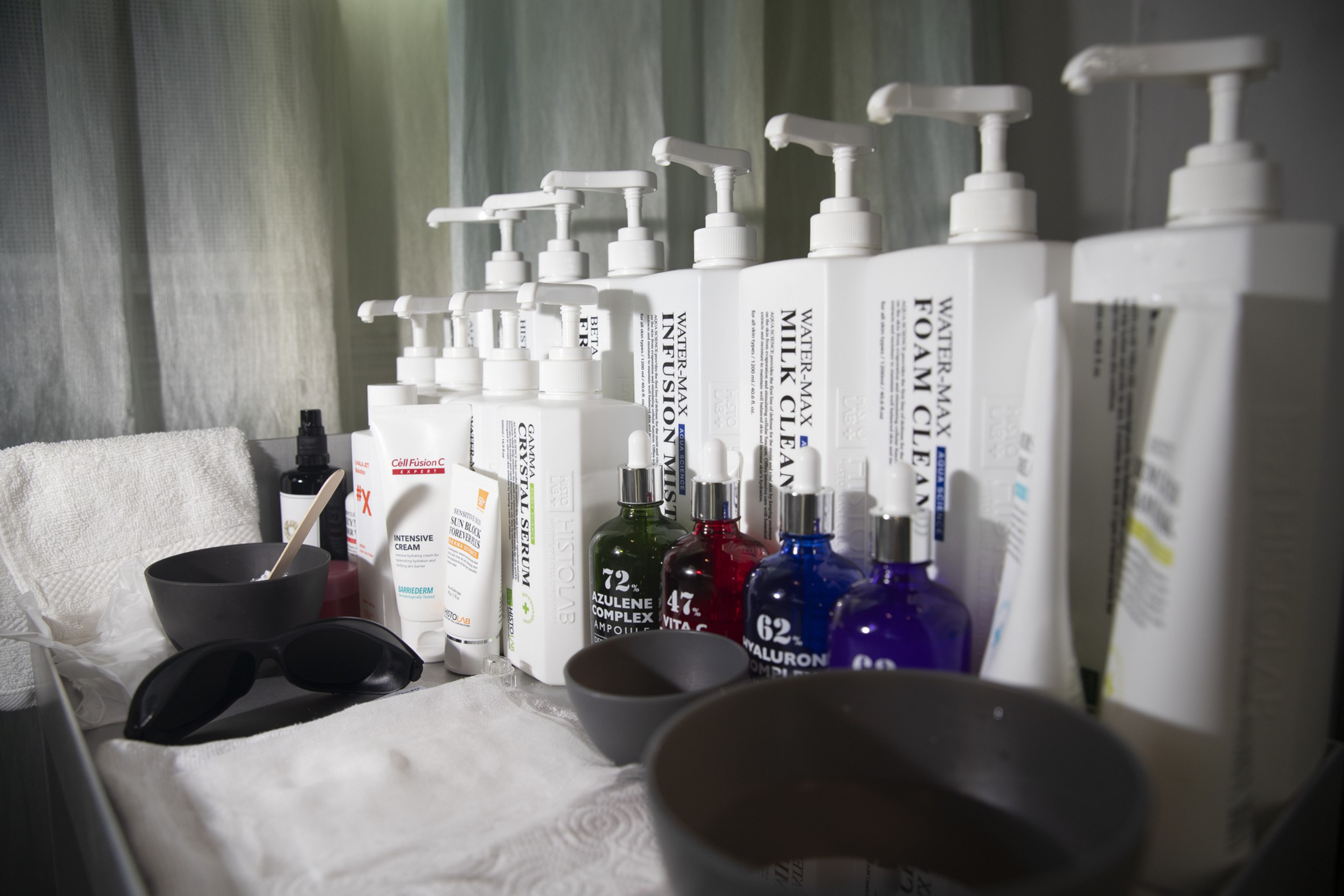 A row of white pump bottles, colorful dropper bottles, skincare tubes, black sunglasses, a white towel, and two dark mixing bowls are arranged on a surface.