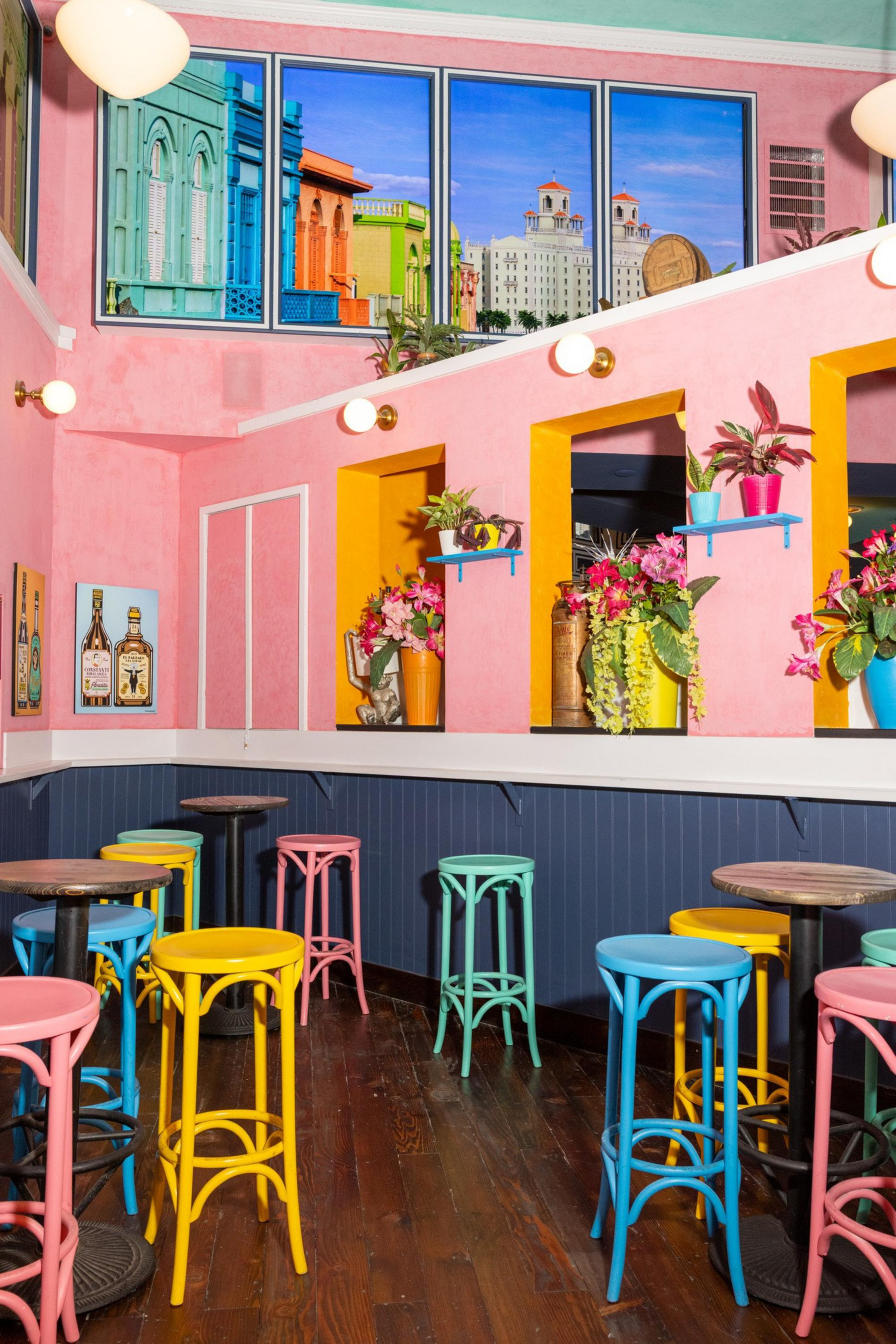 Colorful bar stools in pastel pink, yellow, blue, and green surround wooden tables in a pink-walled room with bright flower pots and architectural artwork.
