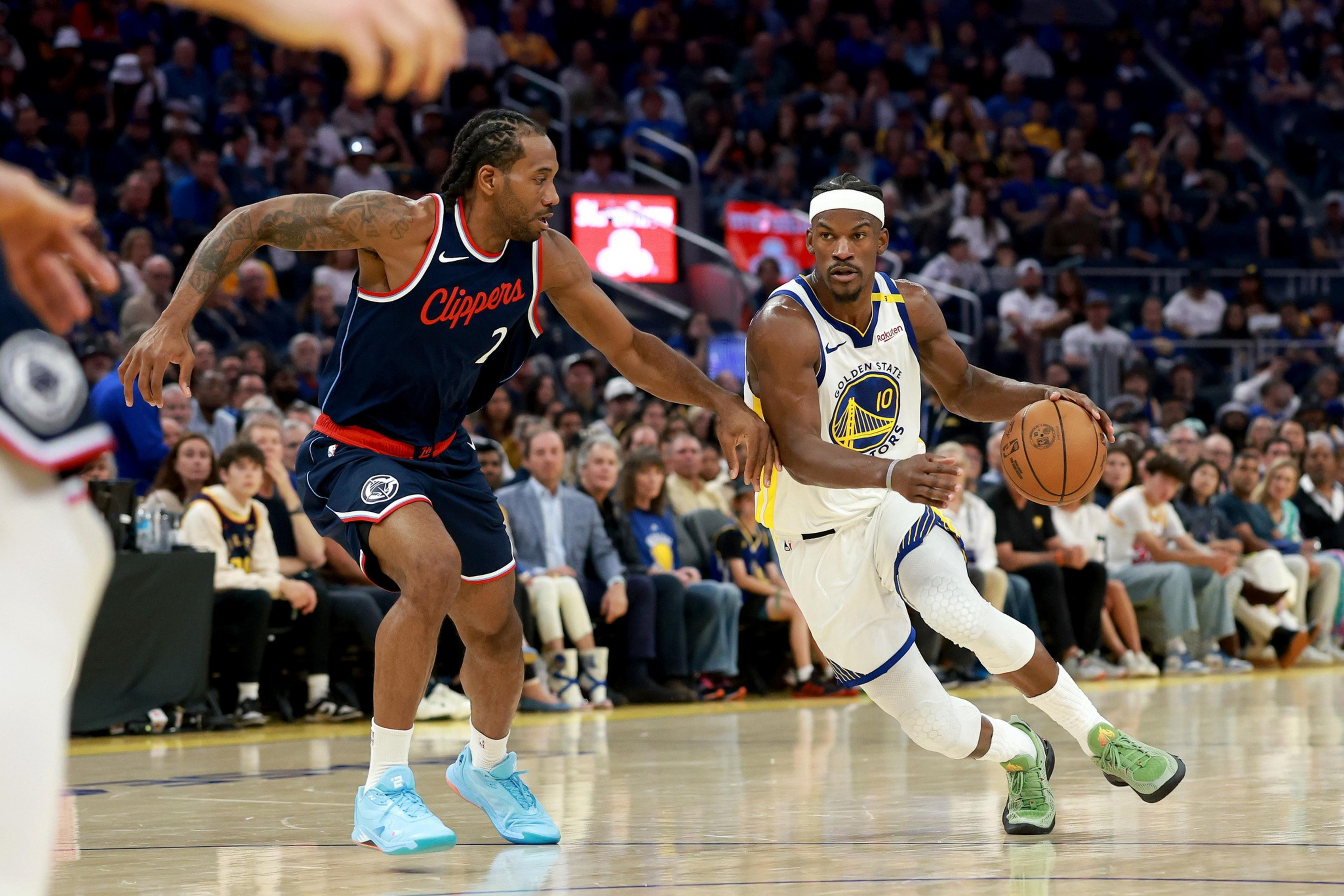 A basketball player from the Golden State Warriors dribbles the ball while a Los Angeles Clippers player closely defends, both surrounded by a crowded arena.