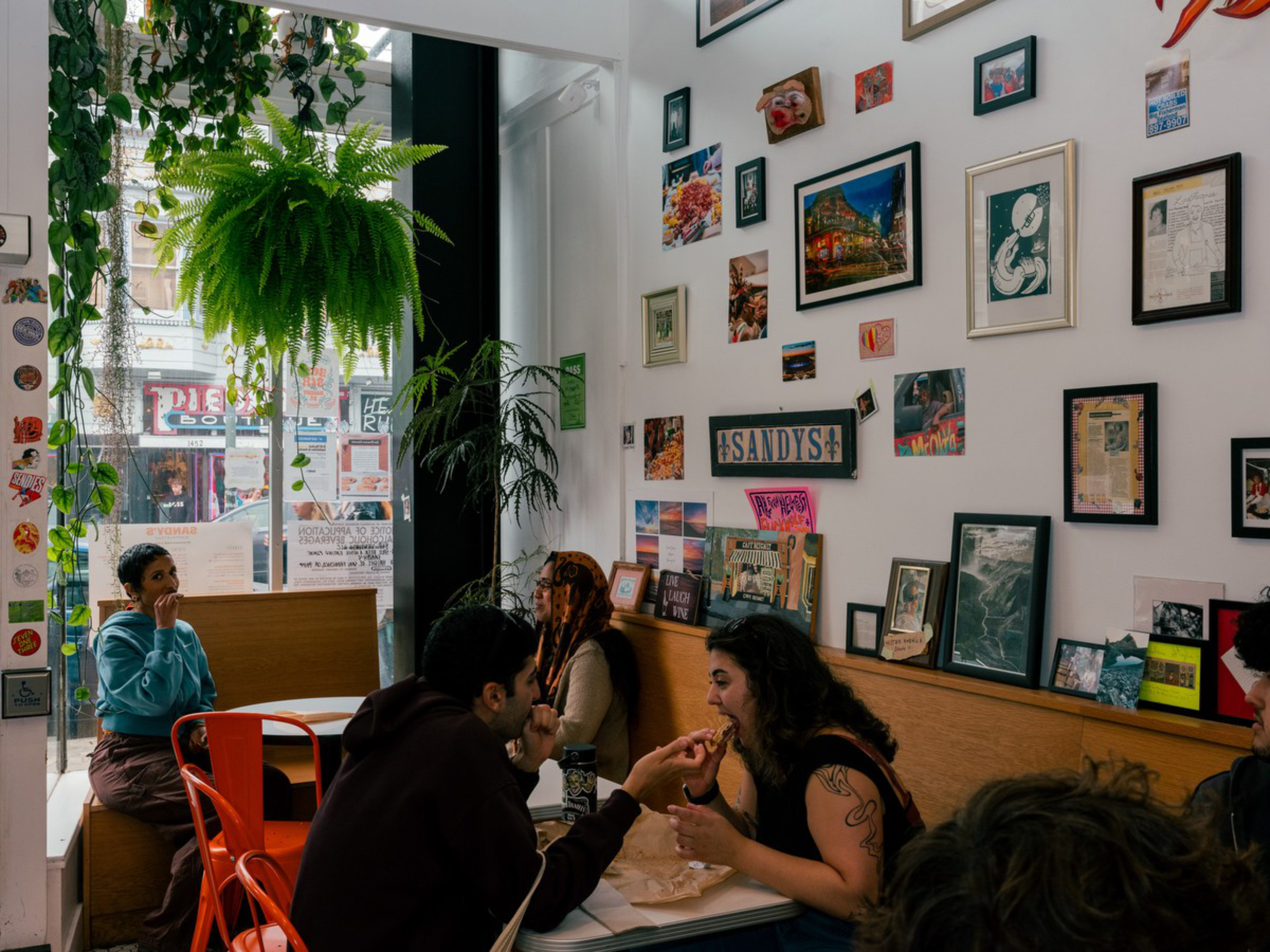 Several people are seated at tables in a cozy cafe with framed pictures and plants, including a large hanging fern, near a window showing street signs.