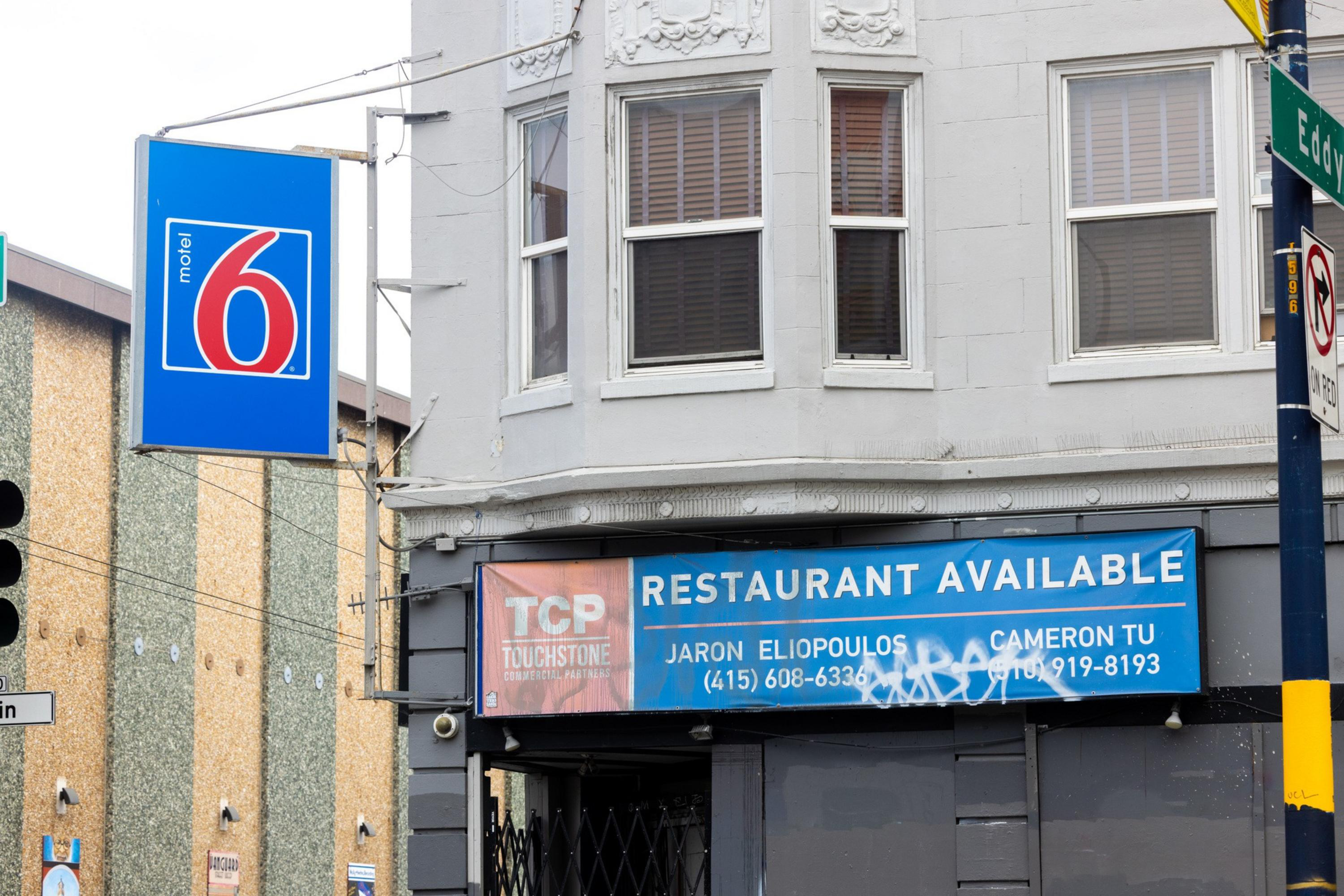 A gray building corner has a blue "Restaurant Available" sign with contact info and a nearby blue Motel 6 sign. Windows are above the signs.