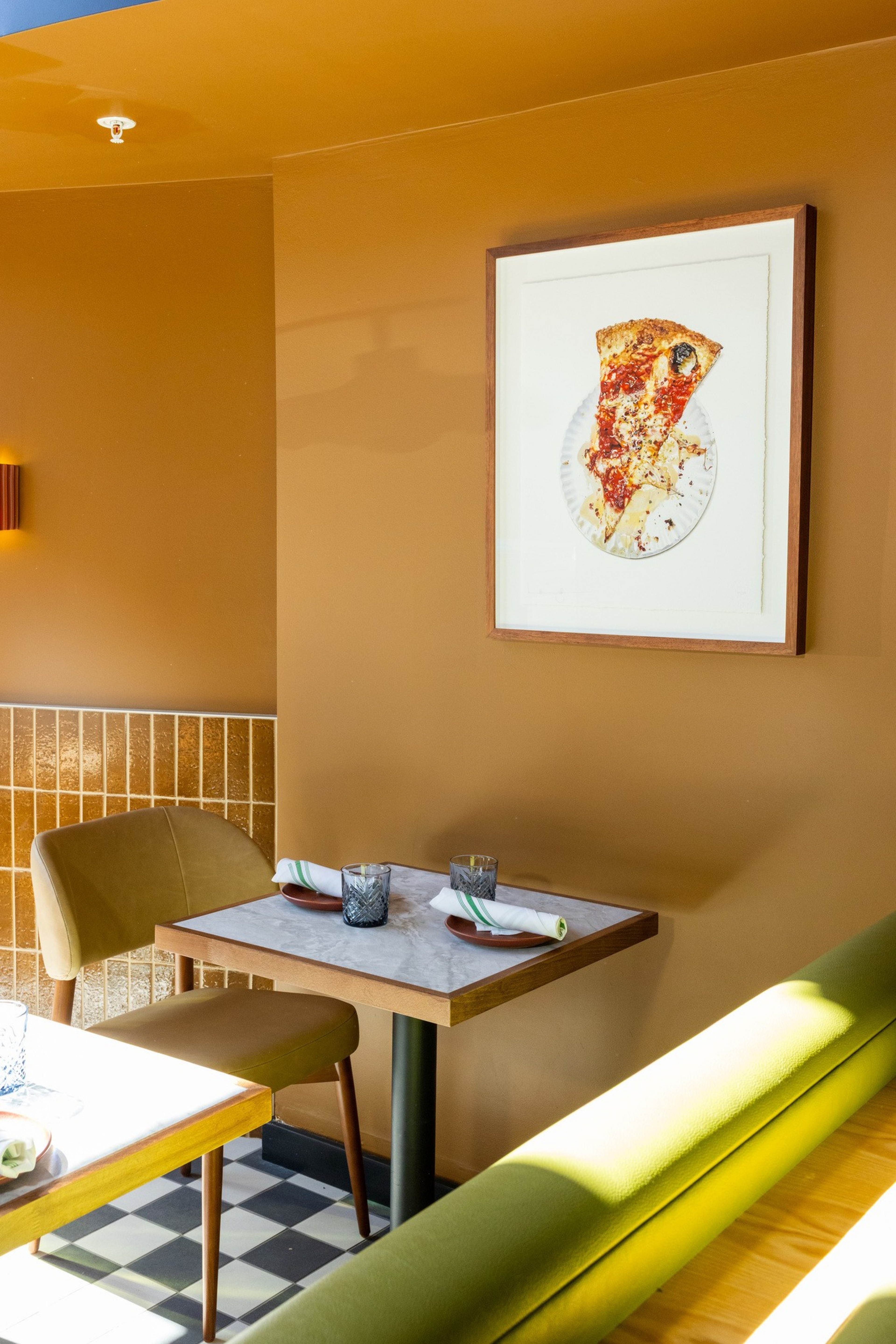 A small dining table with two chairs and two place settings sits against a tan wall with a framed picture of a pizza slice above it.