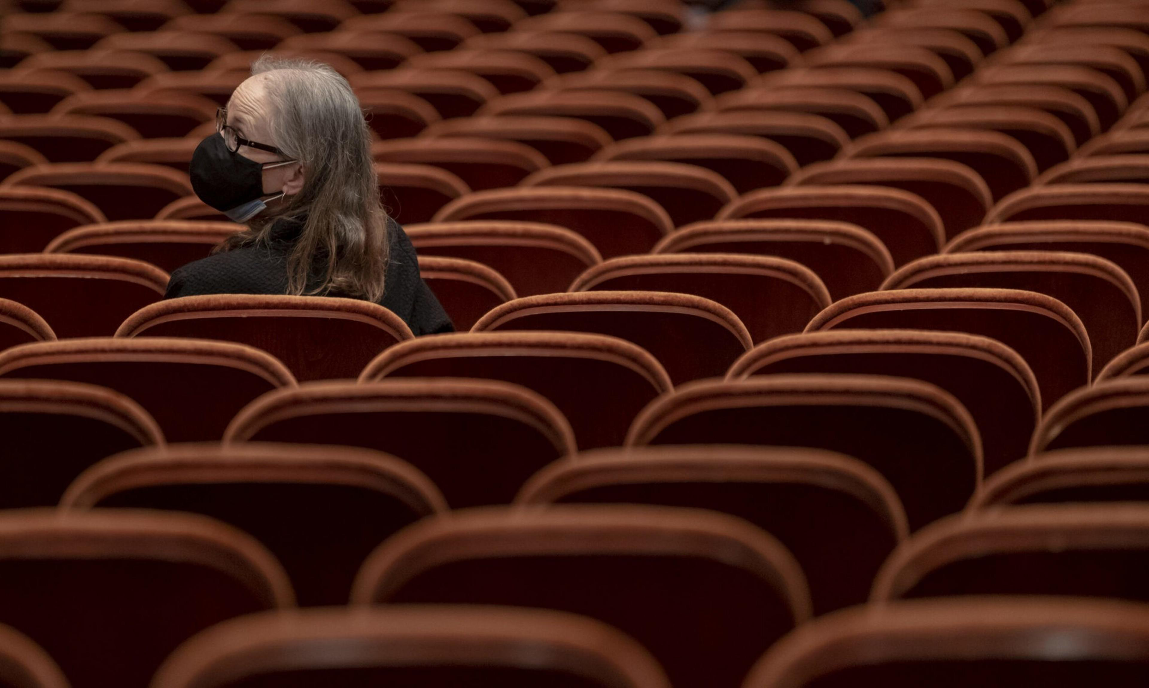 A person with gray hair and glasses wearing a black face mask sits alone, turned to look back, among many empty reddish-brown theater seats.