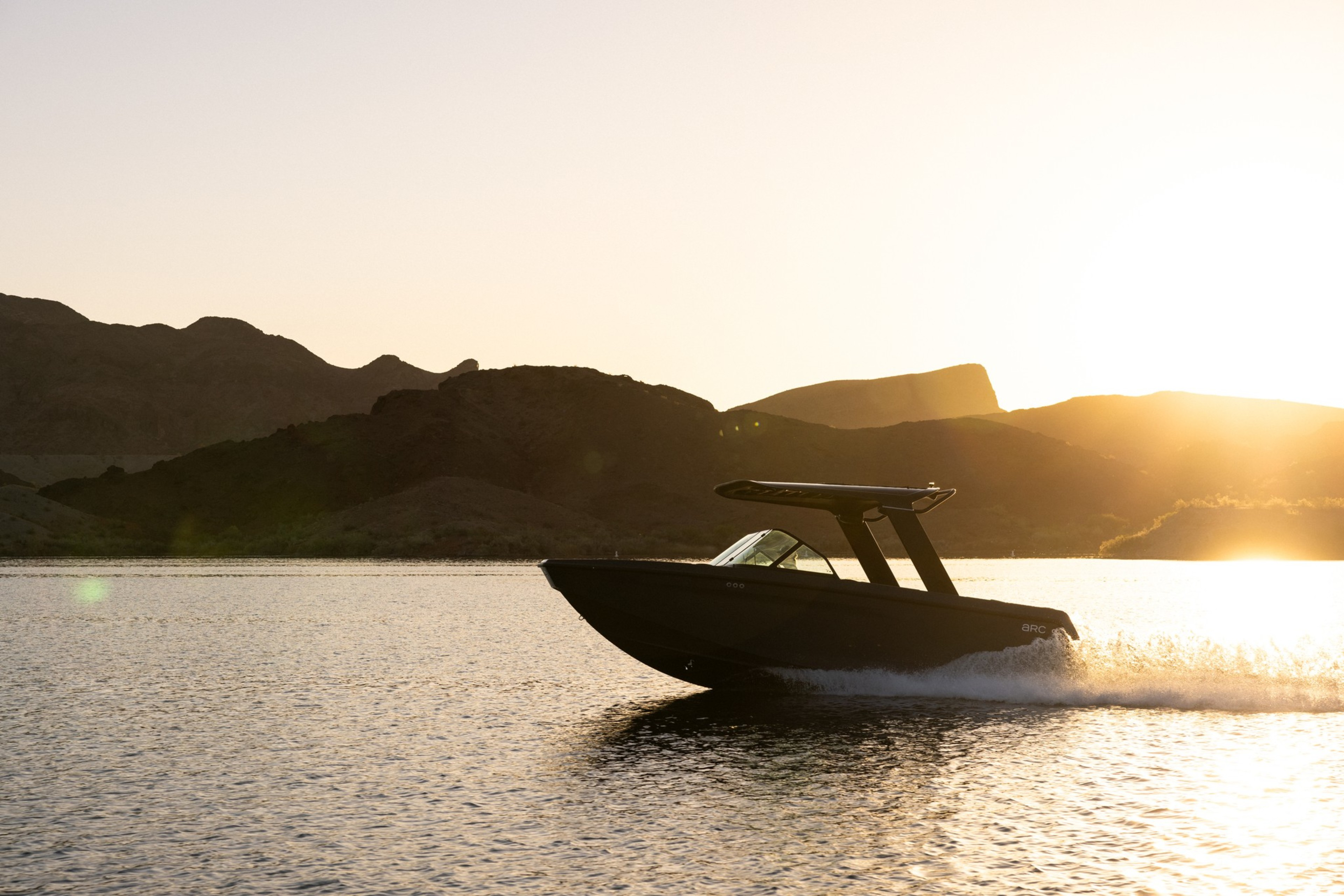 A sleek black boat speeds across a calm lake at sunset, with rugged hills silhouetted in the background and sunlight reflecting off the water.