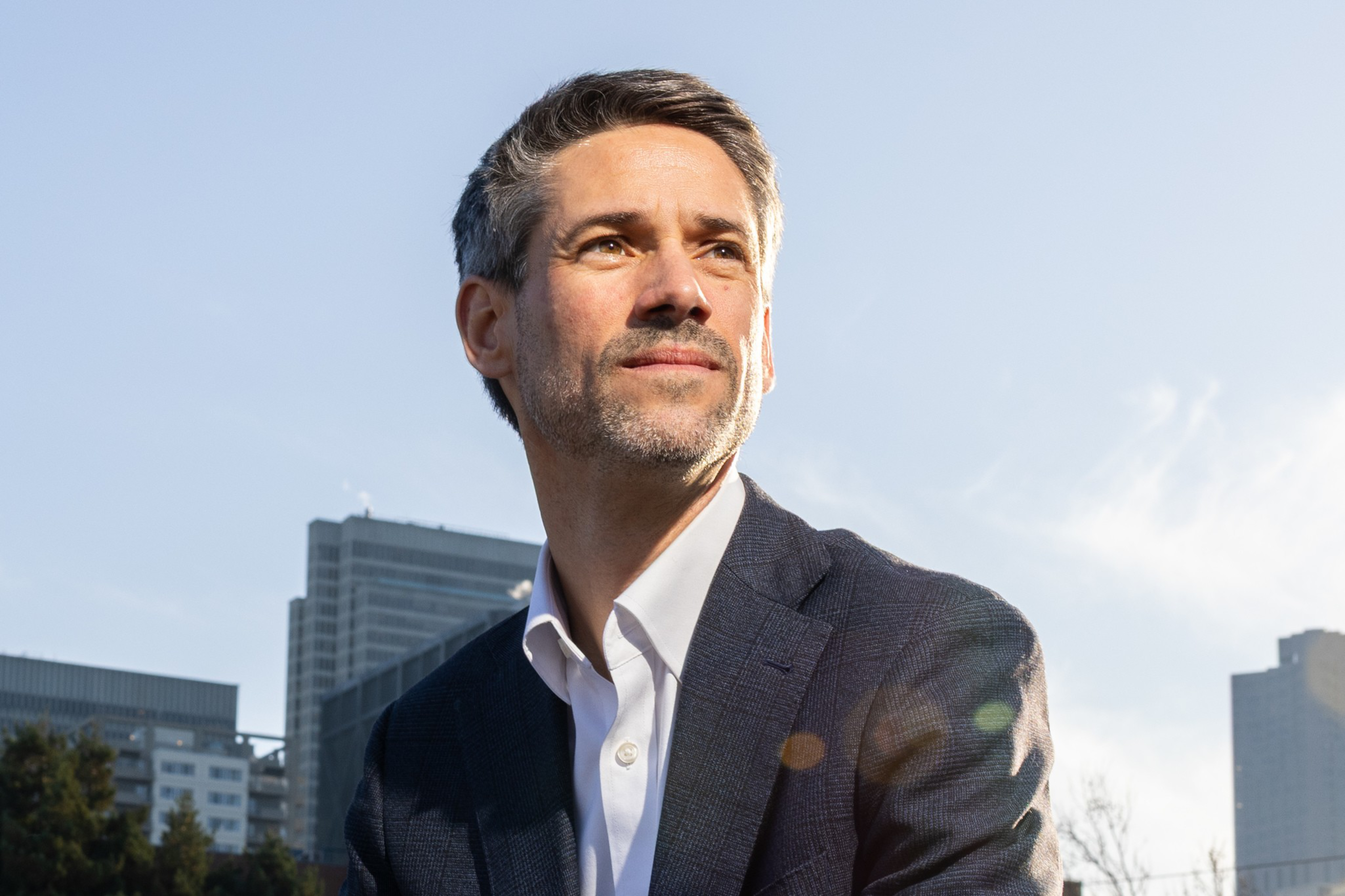 A man in a dark blazer and white shirt looks thoughtfully into the distance with a city skyline and clear sky behind him.