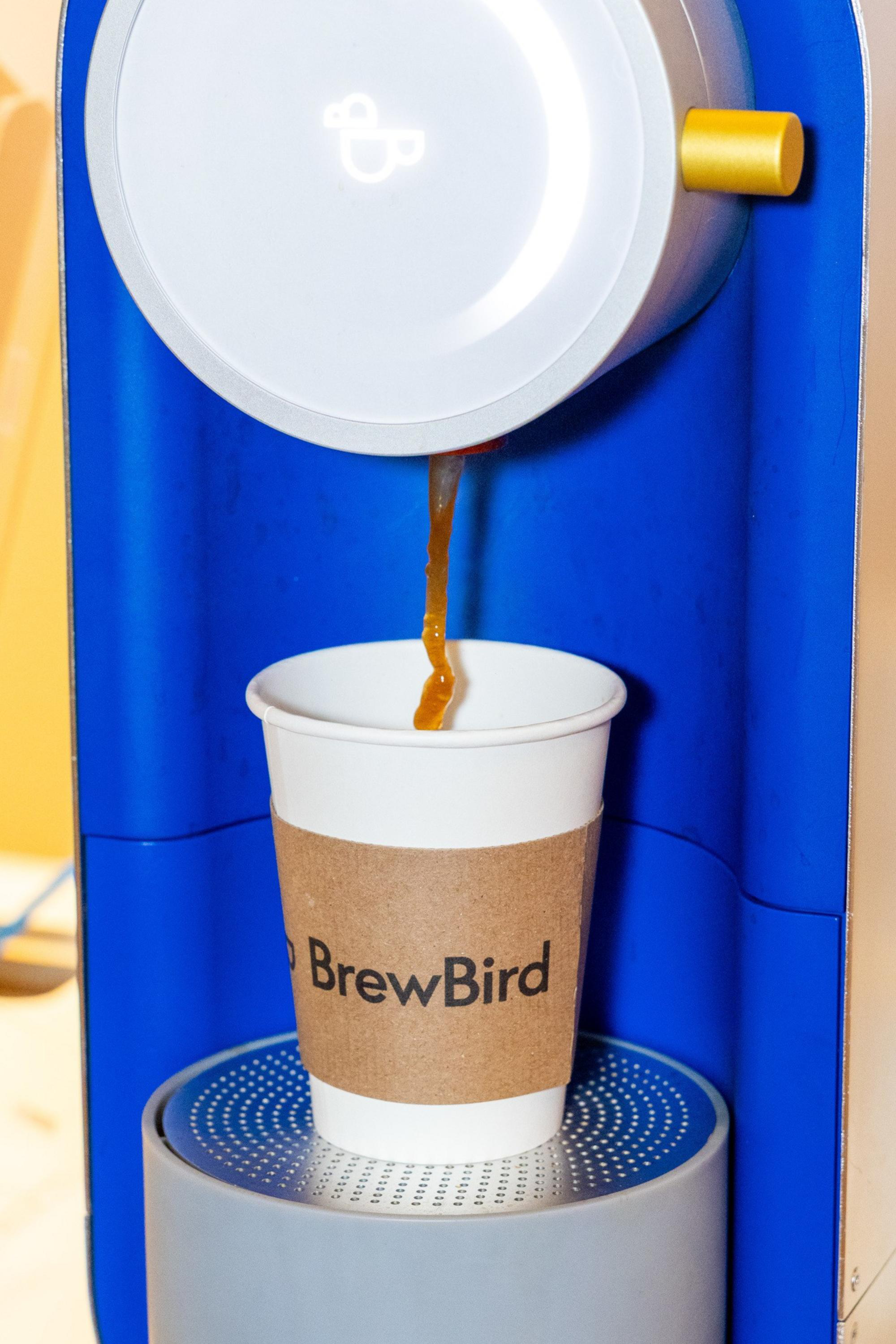 Coffee is flowing from a blue and white BrewBird coffee machine into a paper cup with a brown BrewBird sleeve.