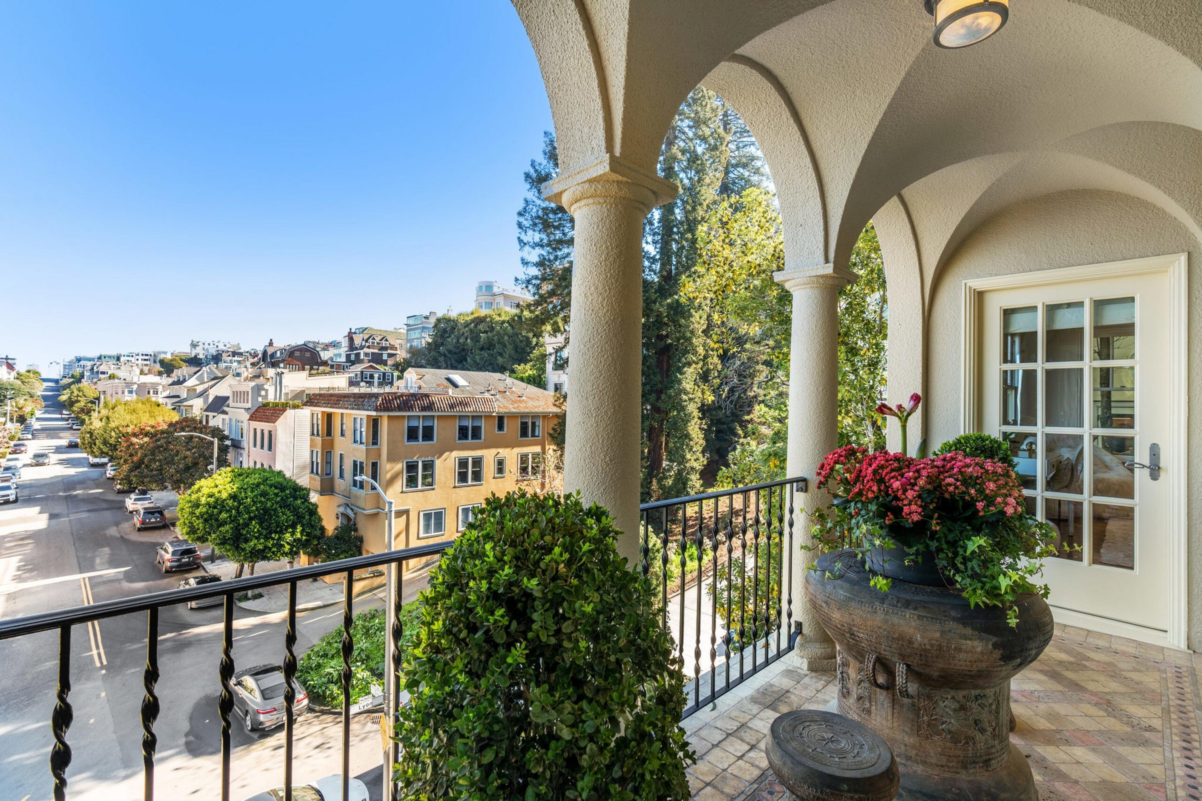 A balcony with arched columns overlooks a tree-lined street, featuring a large stone planter with colorful flowers and a glass-paned door leading indoors.